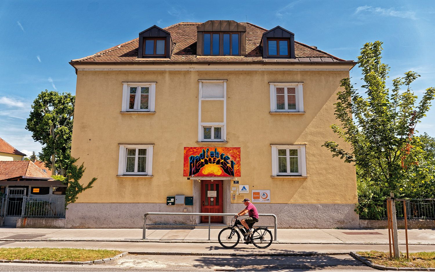 Žlutá budova s červeným vchodem a nápisem graffiti. Kolem projíždí cyklista. V pozadí modrá obloha a stromy.