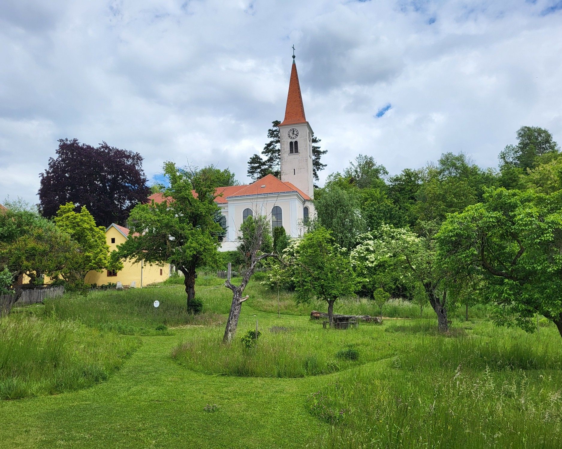 Farní zahrada v Königsbrunnu am Wagram s výhledem na historický kostel, obklopená zelenými loukami, starými ovocnými stromy a přírodní zahradní krajinou.