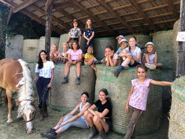 Skupina dětí sedí ve stáji na balících sena a vedle nich stojí kůň. Děti se usmívají a mají na sobě neformální oblečení.