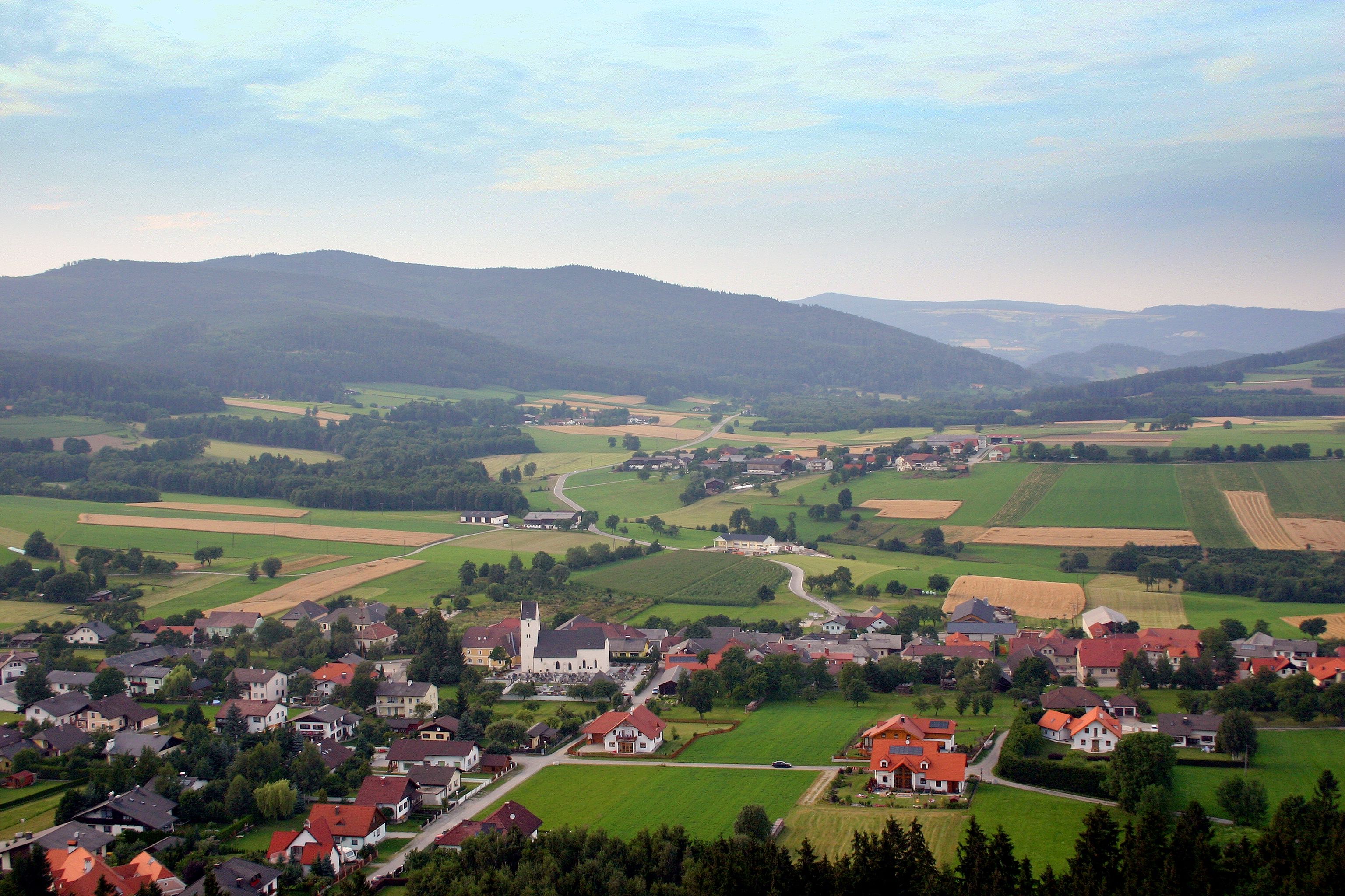 Pohled na krajinu Münichreith am Ostrong s poli, domy a kopci v pozadí.