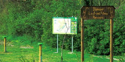 Dřevěná cedule s nápisem "Blumauer Lauf- und Fitness Parcours" před informační tabulí v přírodě.