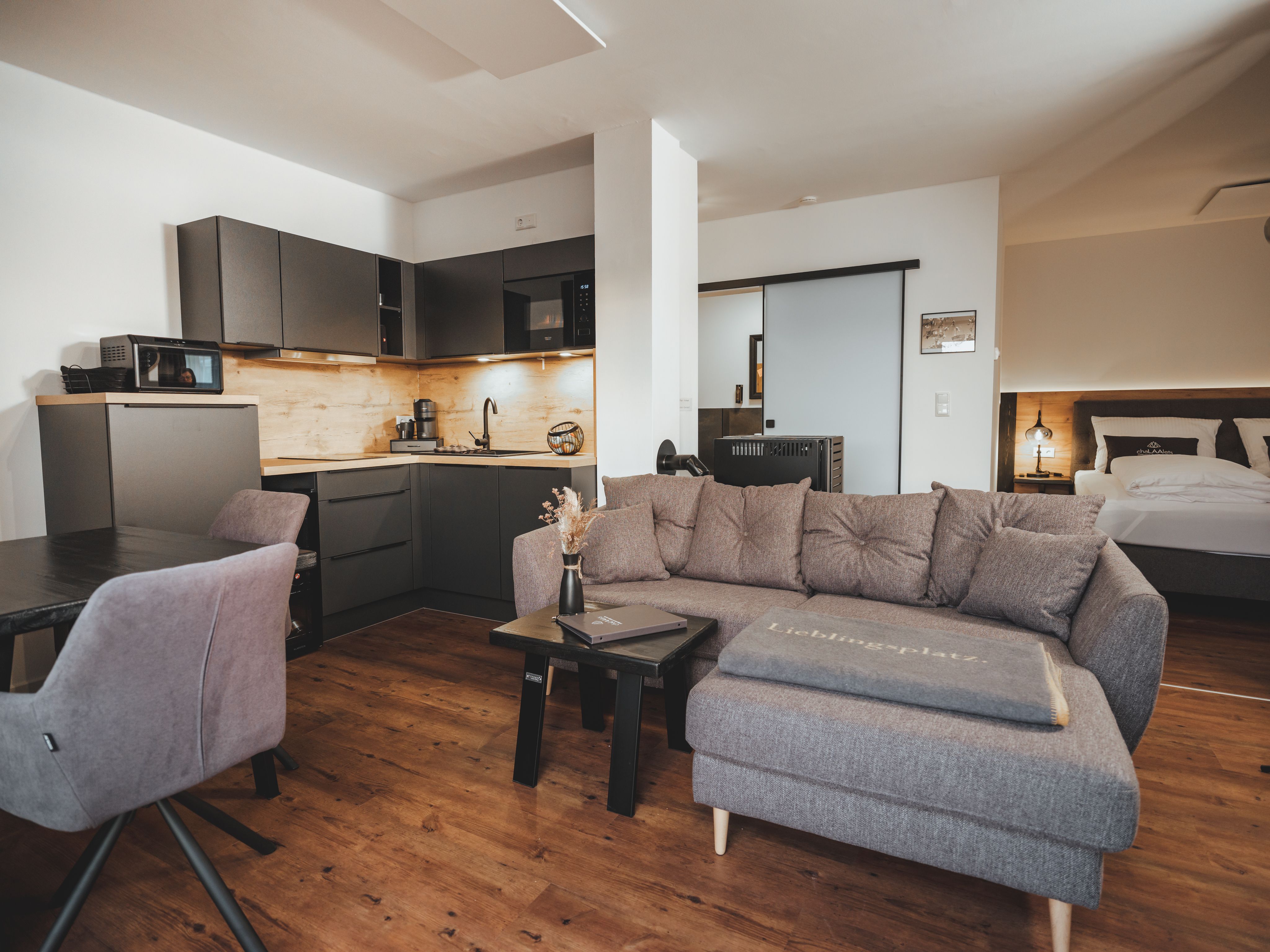 Moderní obývací prostor s šedou pohovkou, kuchyňským koutem a jídelním stolem.