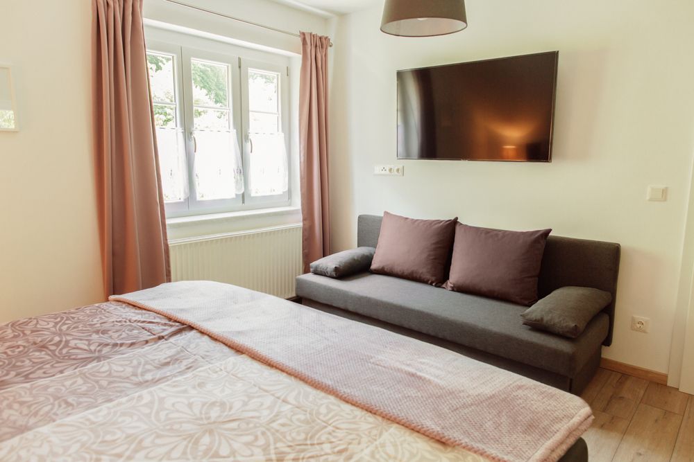 Pohled do interiéru ložnice s postelí, pohovkou, oknem a nástěnnou televizí.