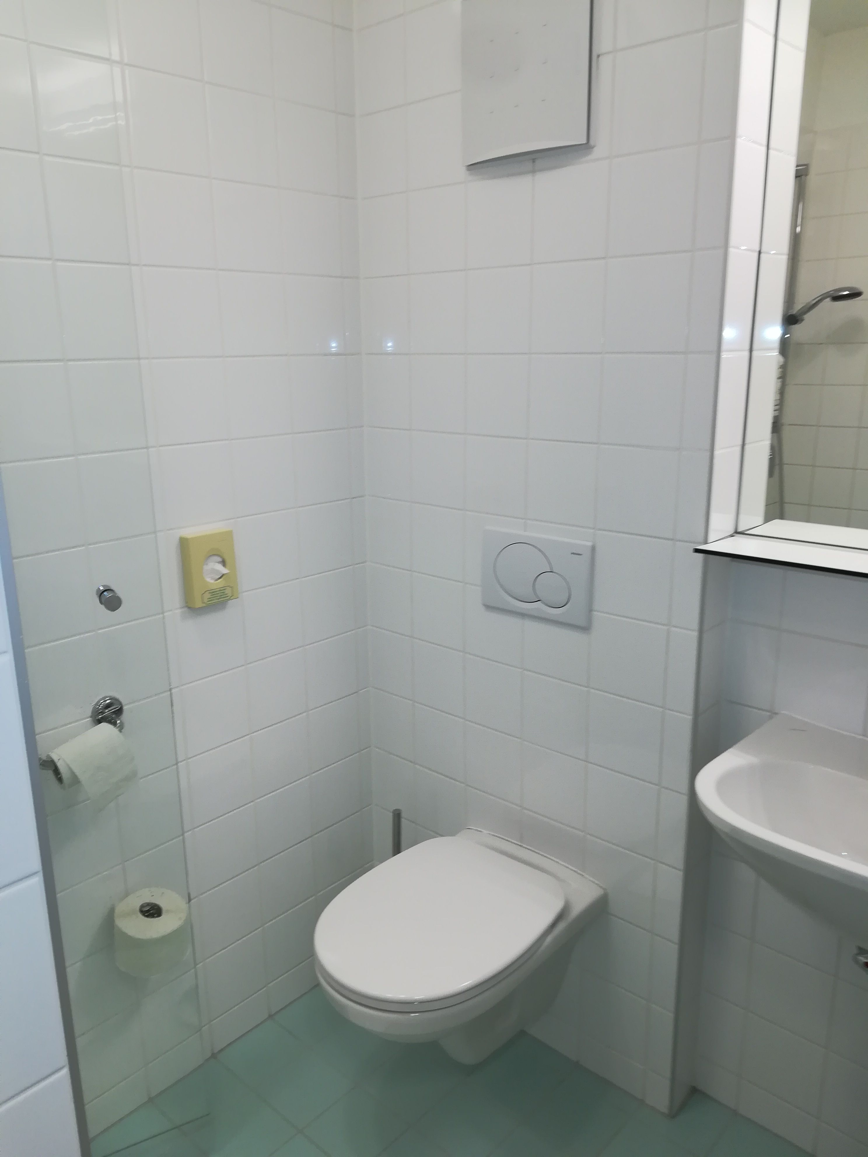 Malá koupelna s bílými kachličkami, toaletou, umyvadlem a držákem na toaletní papír.