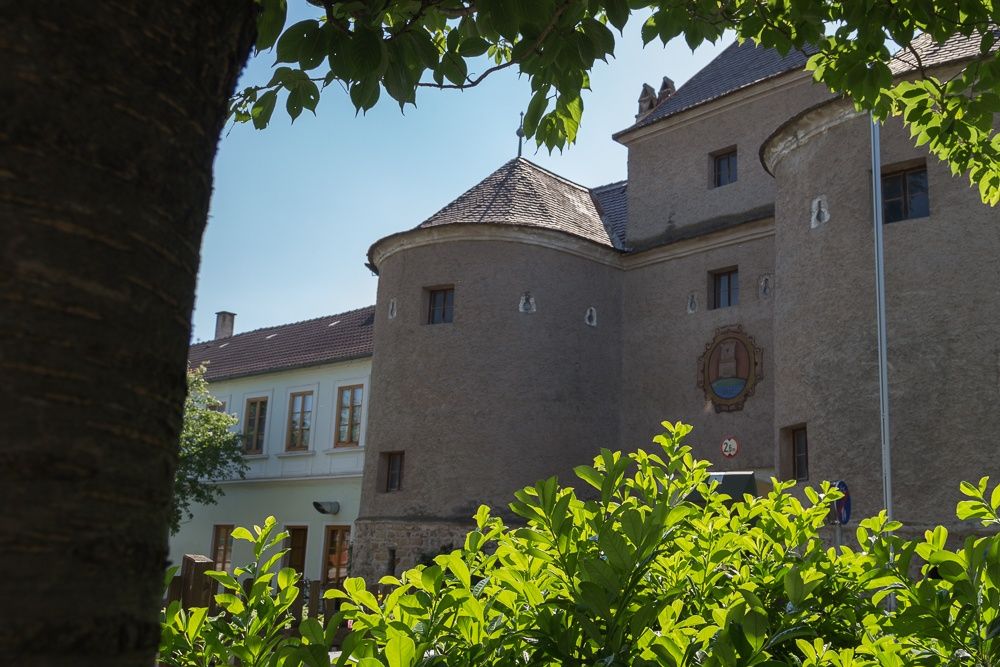 Historická budova s kulatými věžemi a erbem, obklopená zelenými stromy a keři.