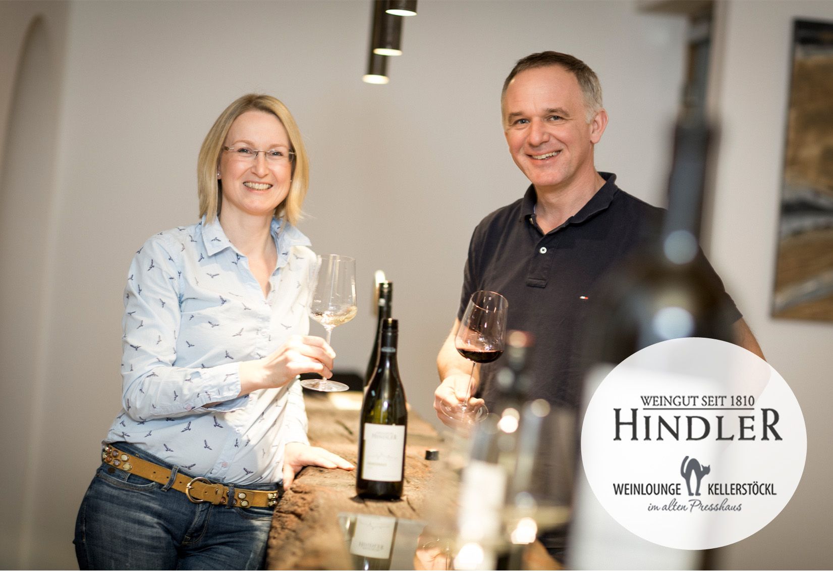 Dvojice vinařů stojí s úsměvem a skleničkami v rukou u stolu ve vinařství Hindler.