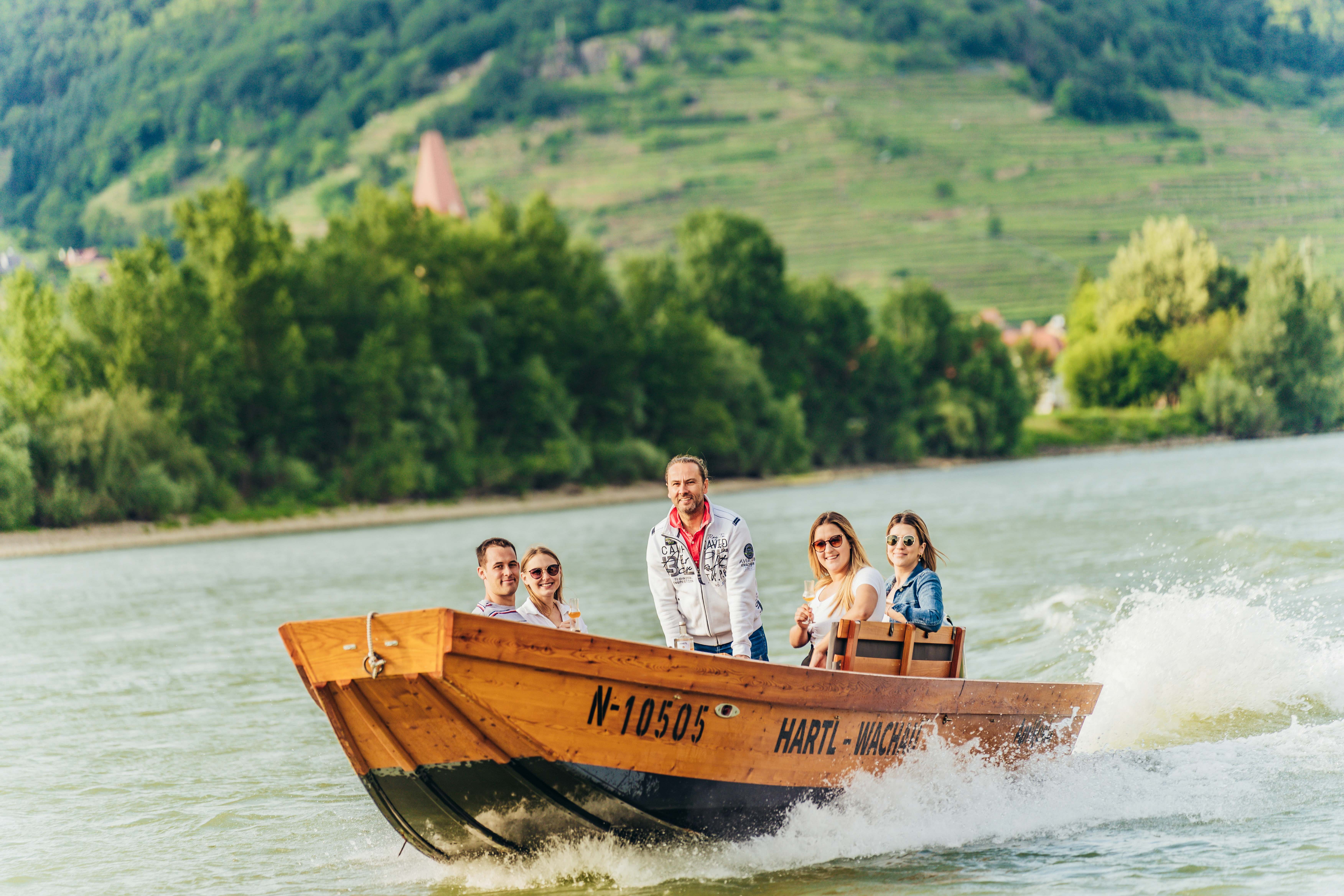 Skupina lidí na dřevěné lodi na Dunaji se zelenou krajinou v pozadí.