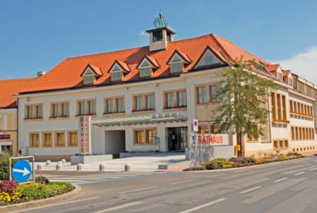 Traiskirchenská radnice s červenou střechou a několika okny, obklopená ulicí a květinovými záhony.