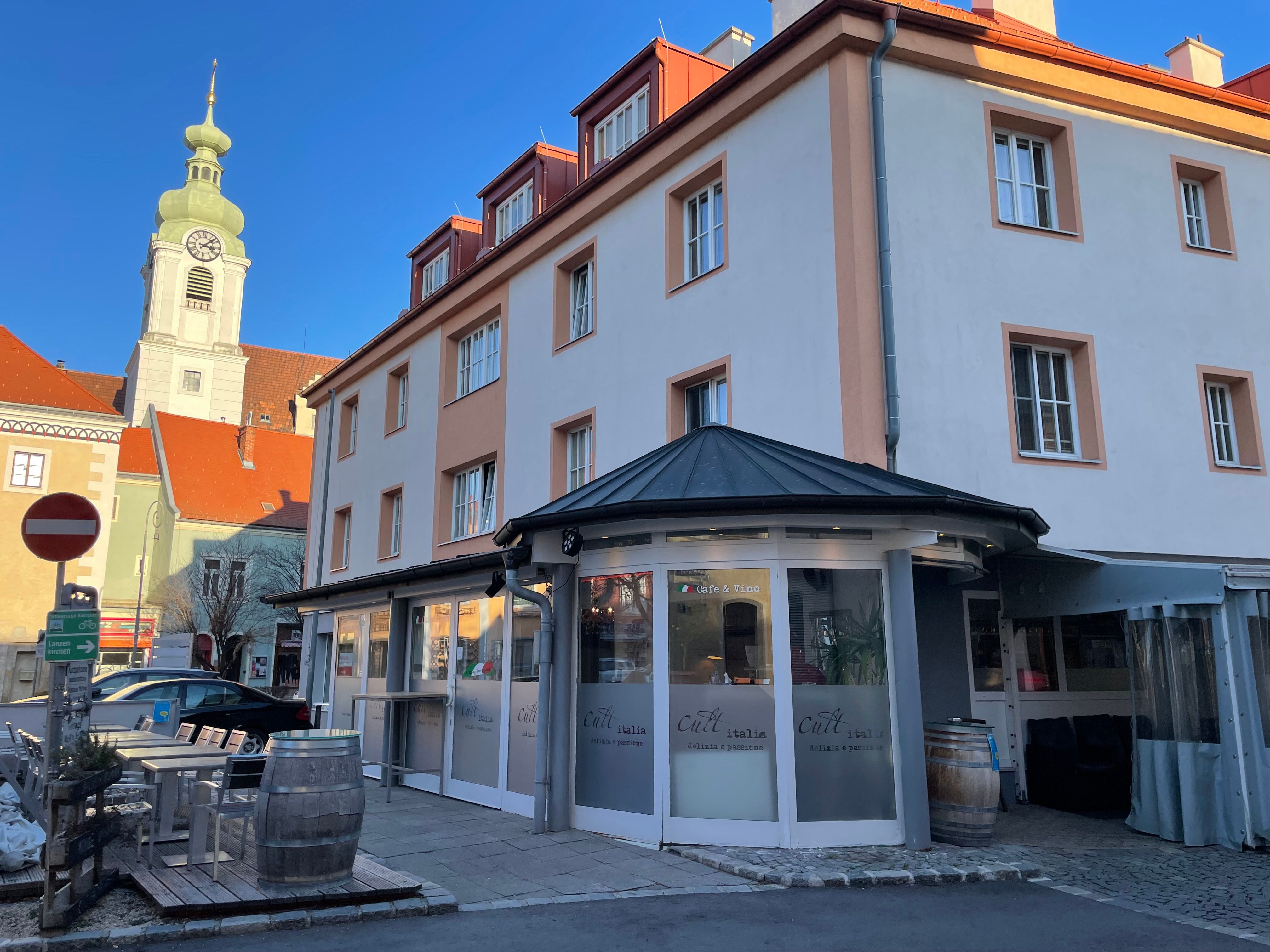 Pohled zvenčí na restauraci s nápisem "Cult Italia" s kostelní věží v pozadí.