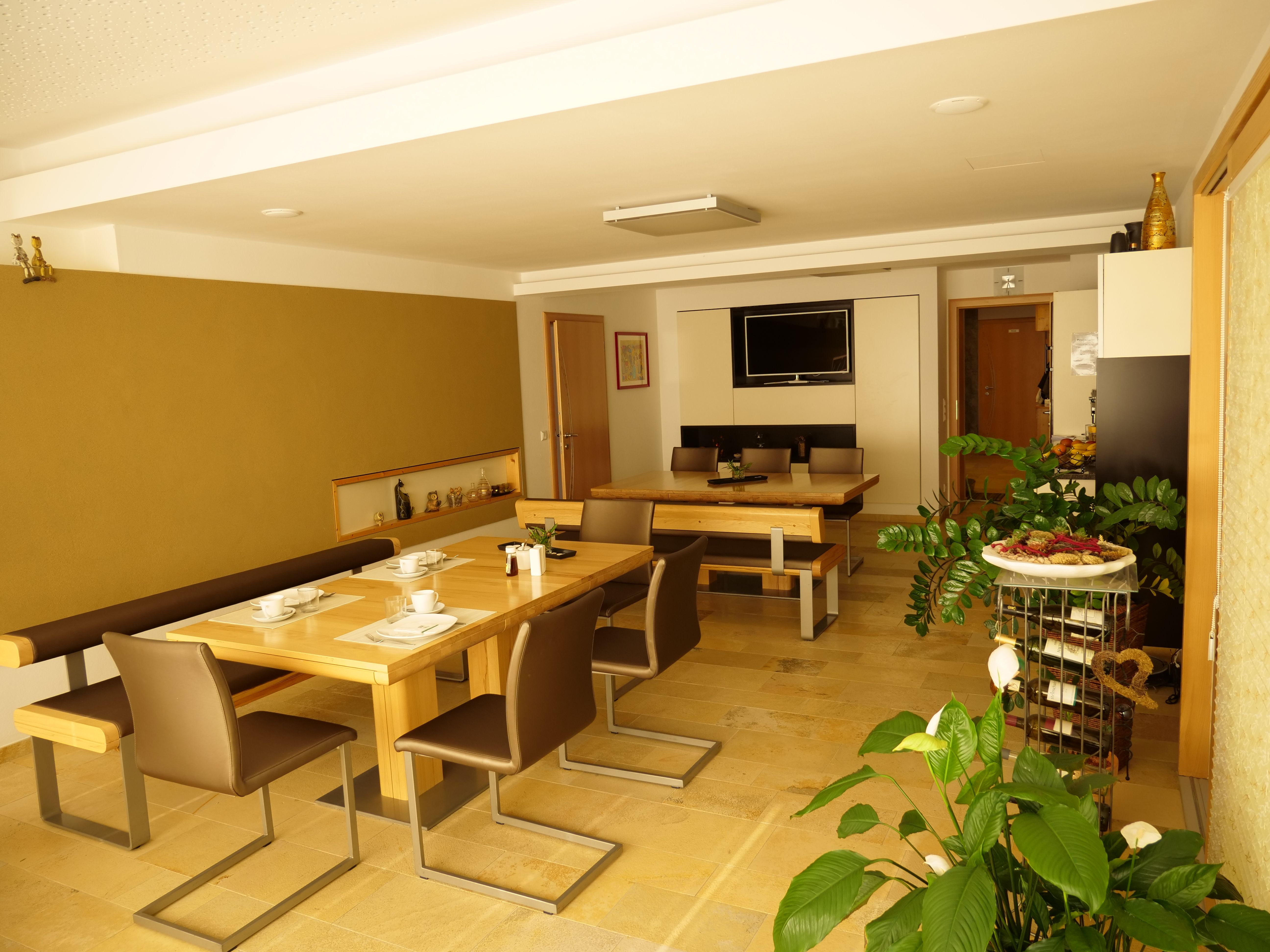 Snídaňová místnost s dřevěnými stoly, židlemi a rostlinami, zdobená nádobím a televizí na stěně.