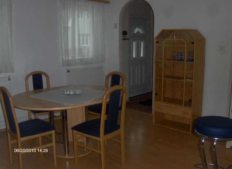 Jídelní kout s kulatým stolem, čtyřmi židlemi a dřevěnou policí ve světlé místnosti.