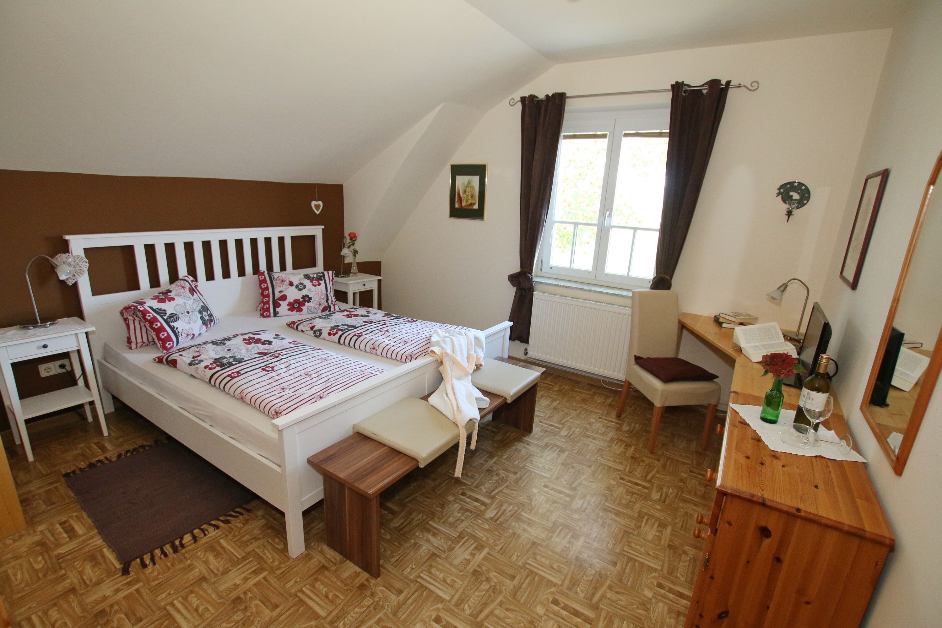 Útulný pokoj pro hosty s manželskou postelí, pohovkou, stolem a židlí. Hnědá akcentní stěna a tmavé závěsy.