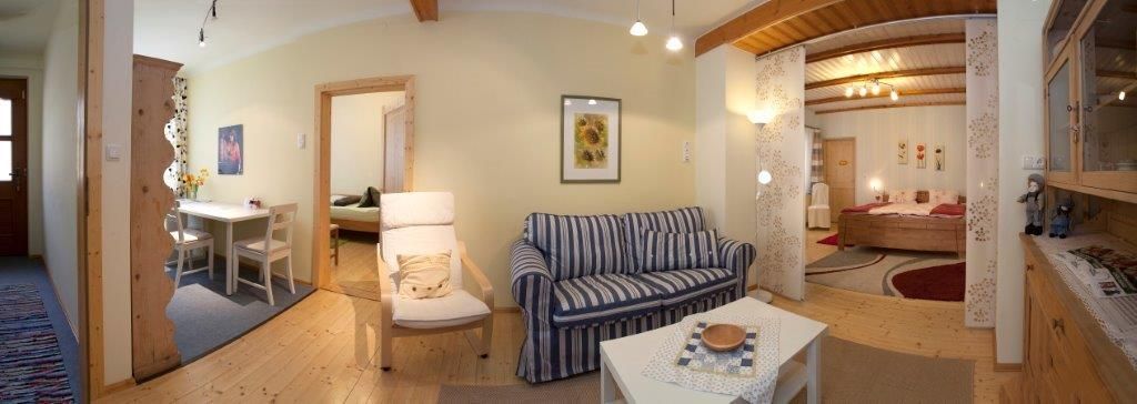 Panorama útulného obývacího pokoje s pohovkou, jídelním stolem a ložnicí v pozadí.