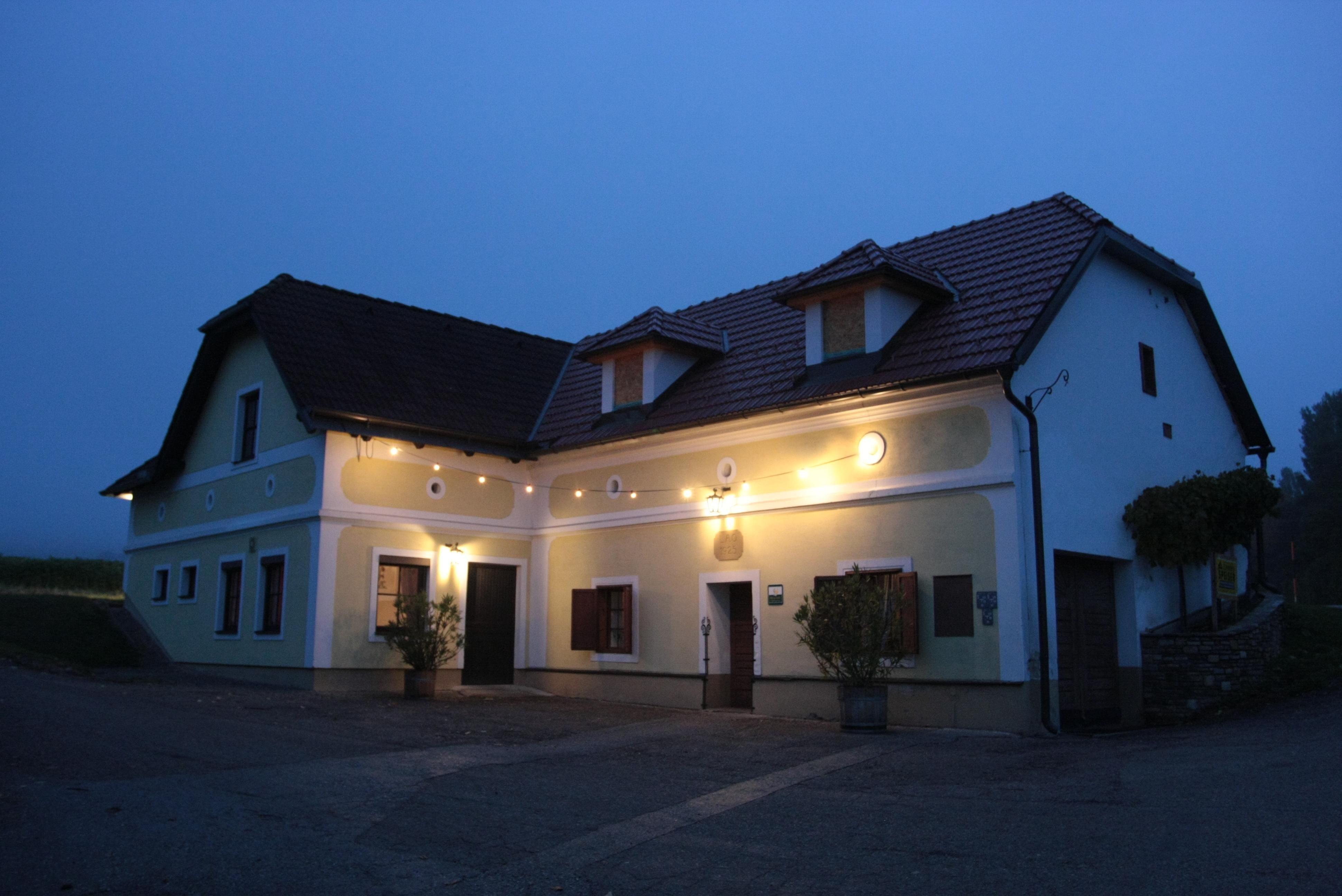 Tradiční osvětlený dům se žlutou fasádou a červenými střešními taškami za soumraku.