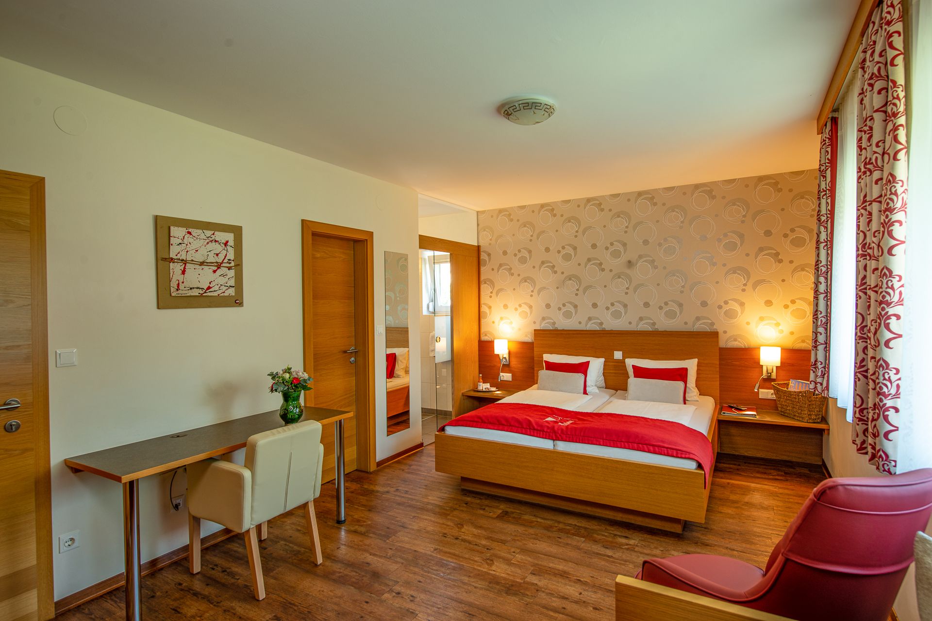 Hotelový pokoj s manželskou postelí, psacím stolem, židlí a červeným křeslem.