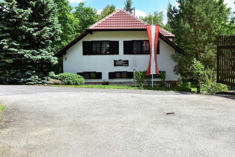 Bílý dům s červenou střechou a rakouskou vlajkou před ním, obklopený stromy.