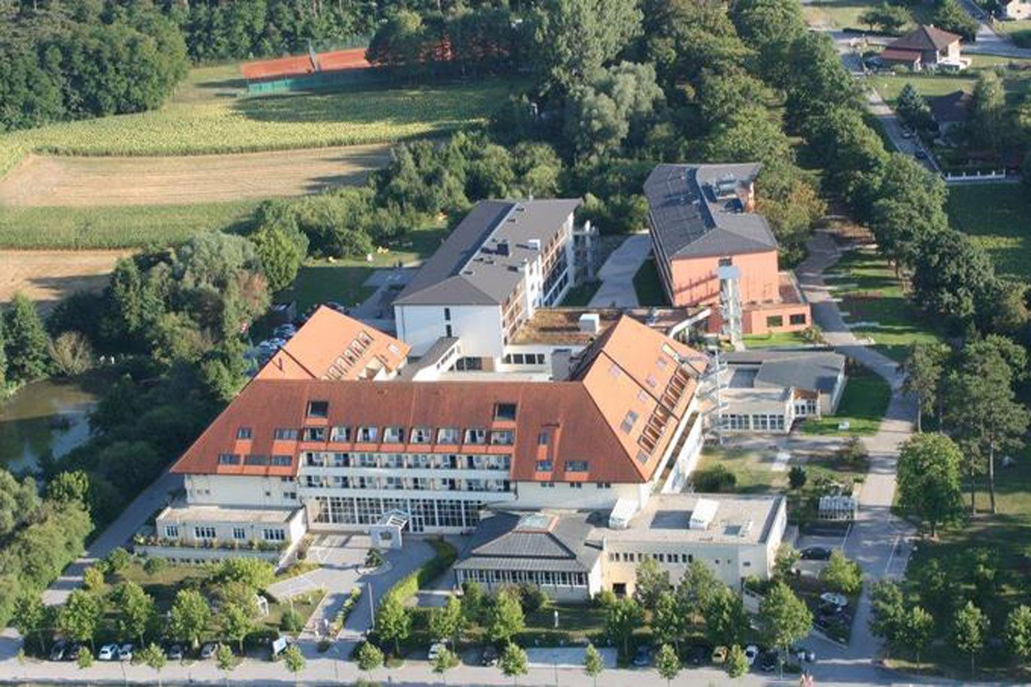Letecký pohled na kliniku s červenými střechami, obklopenou stromy a poli.