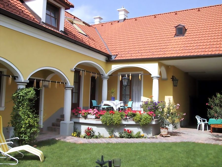 Žlutý dům s červenou střechou, arkádami a terasou s květinami.
