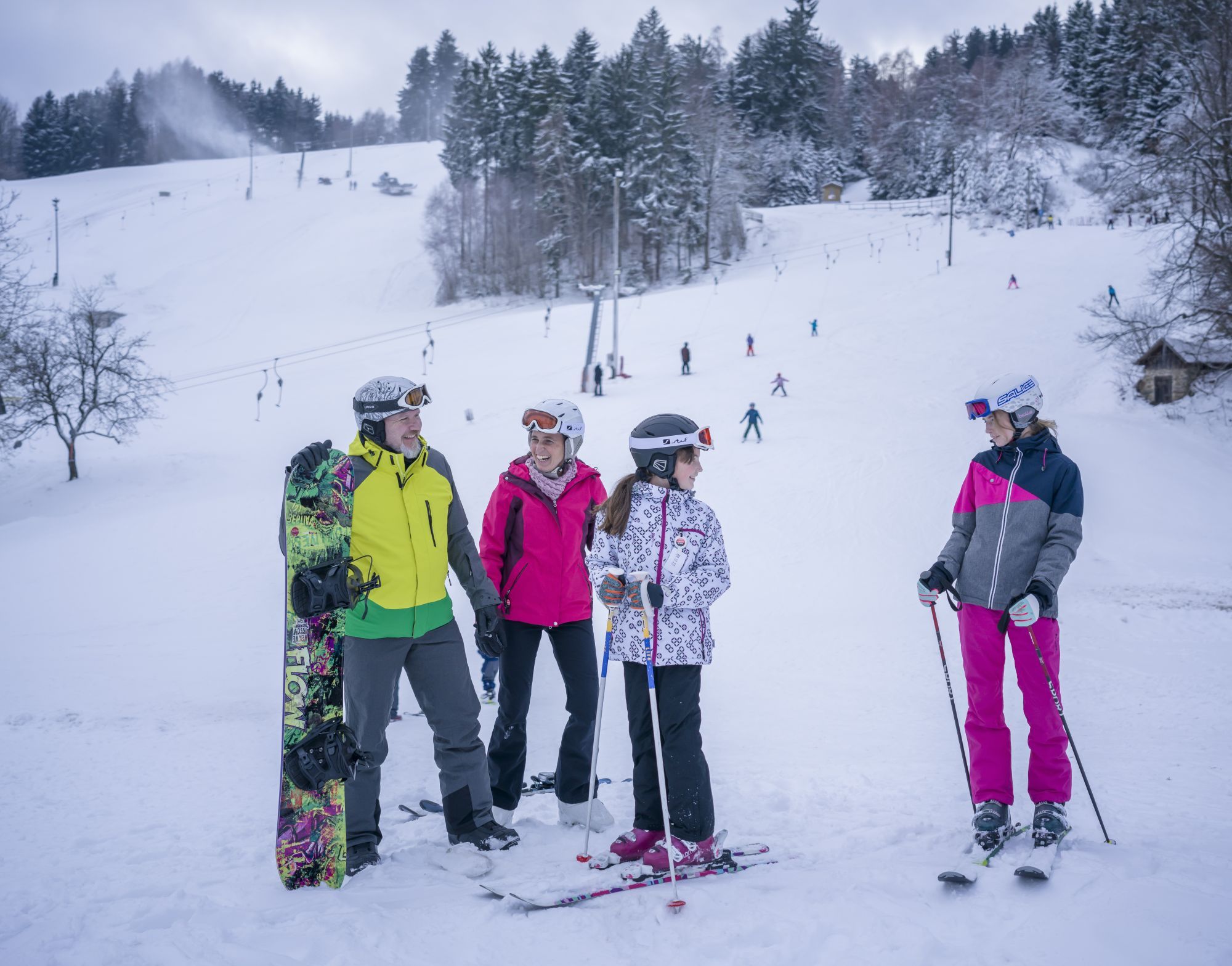 Skupina lyžařů a snowboardistů na sněhu před sjezdovkou s vlekem.
