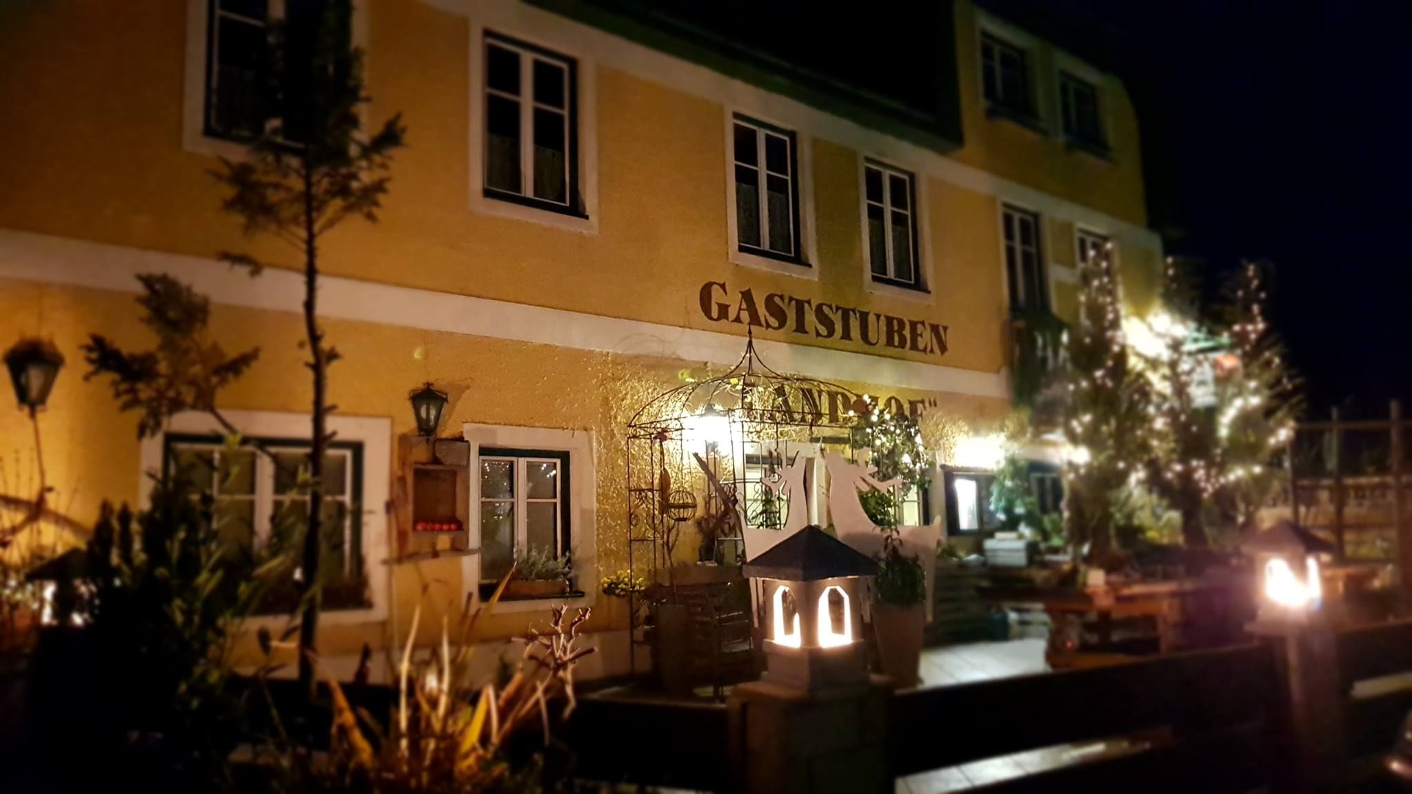 Noční pohled na žlutou budovu s nápisem "Gaststuben Landhof", osvětlenou lucernami a obklopenou rostlinami.