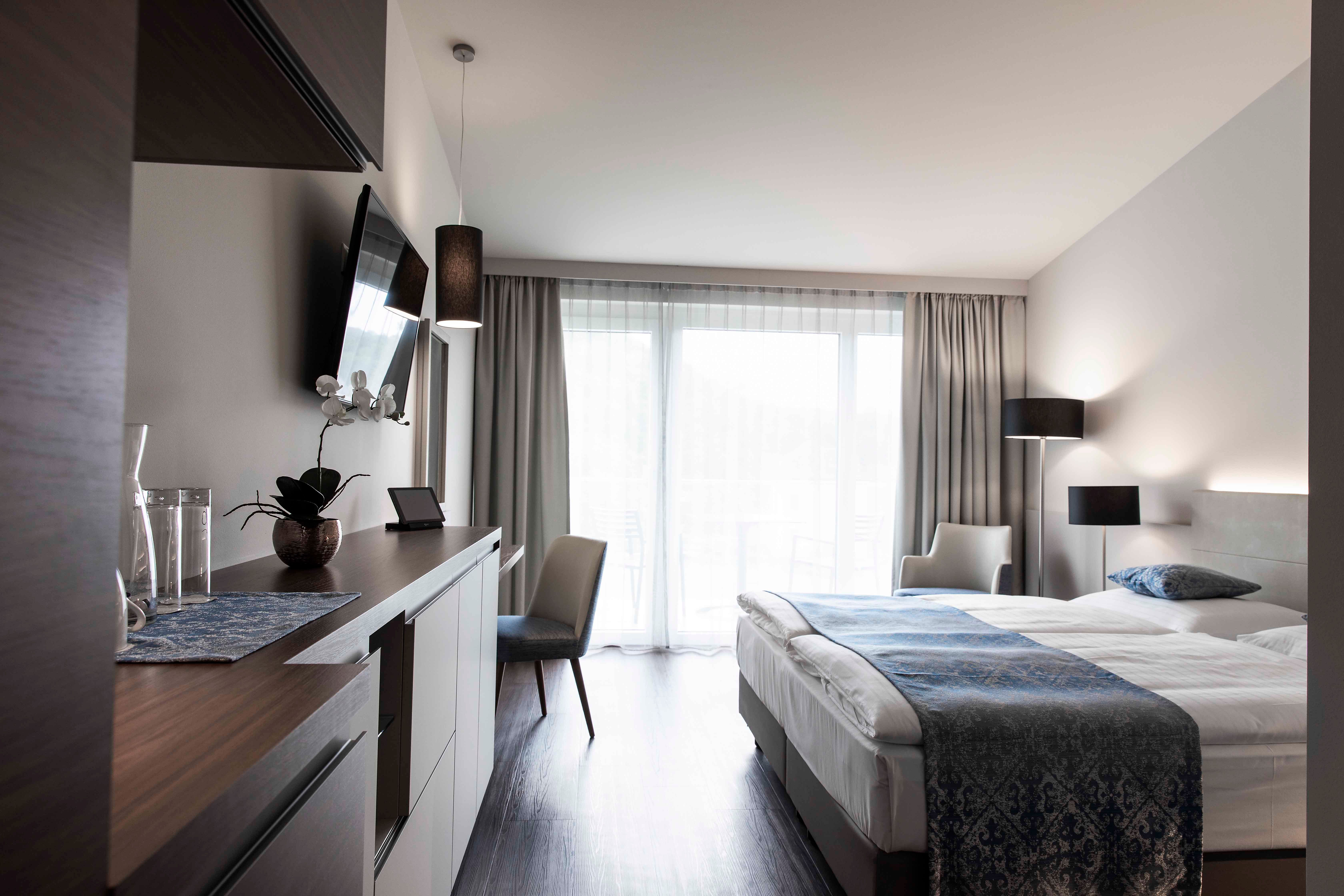 Moderní hotelový pokoj s manželskou postelí, TV, psacím stolem a velkými okny se závěsy.