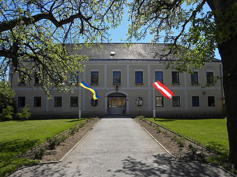 Zámek Hochwolkersdorf se dvěma vlajkami, obklopený stromy a trávníkem.