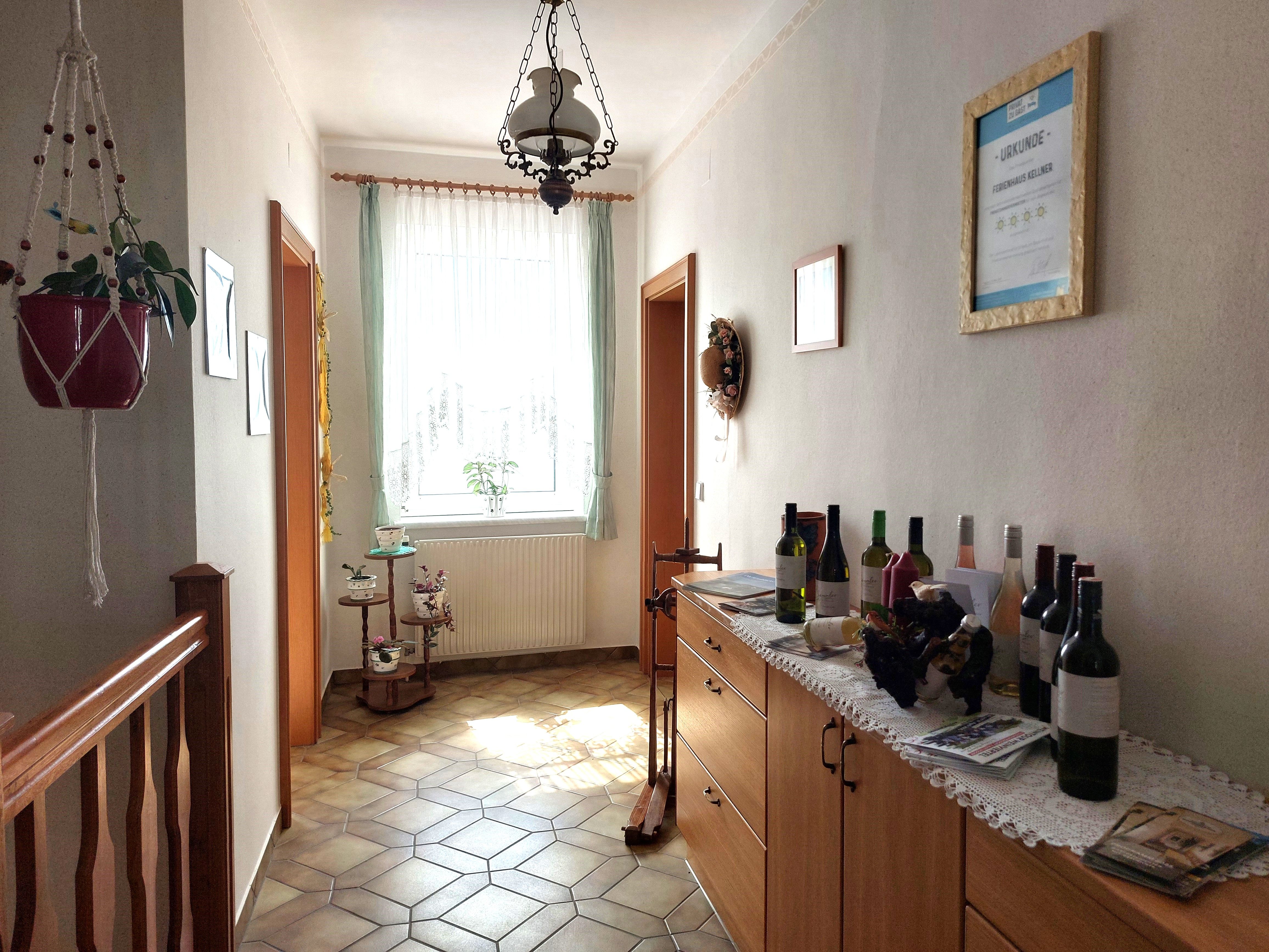 Světlá předsíň s dlážděnou podlahou, komodou s lahvemi vína a oknem se závěsy.