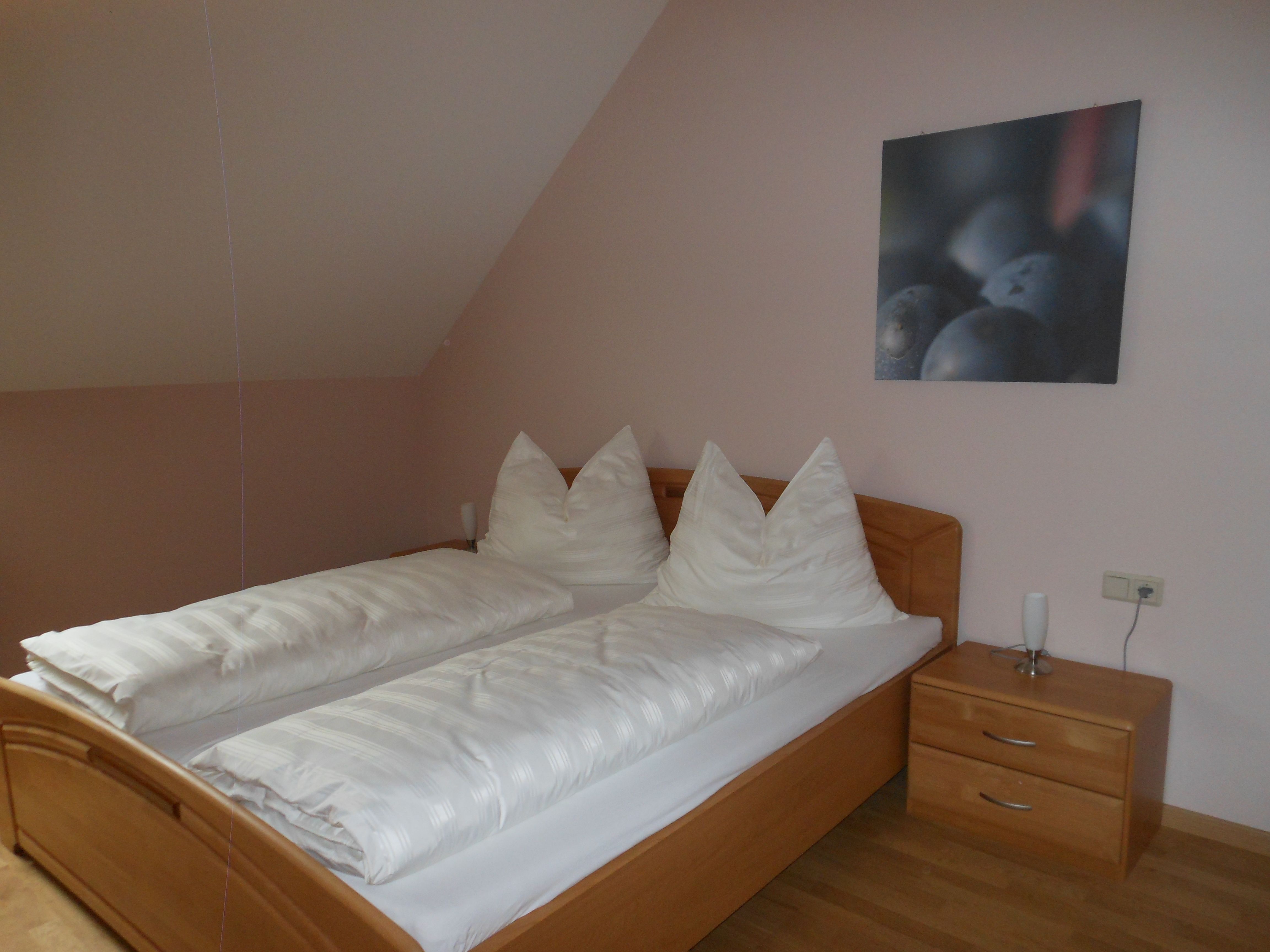 Jednoduchý pokoj pro hosty s manželskou postelí, bílými přikrývkami a nočním stolkem s lampičkou.