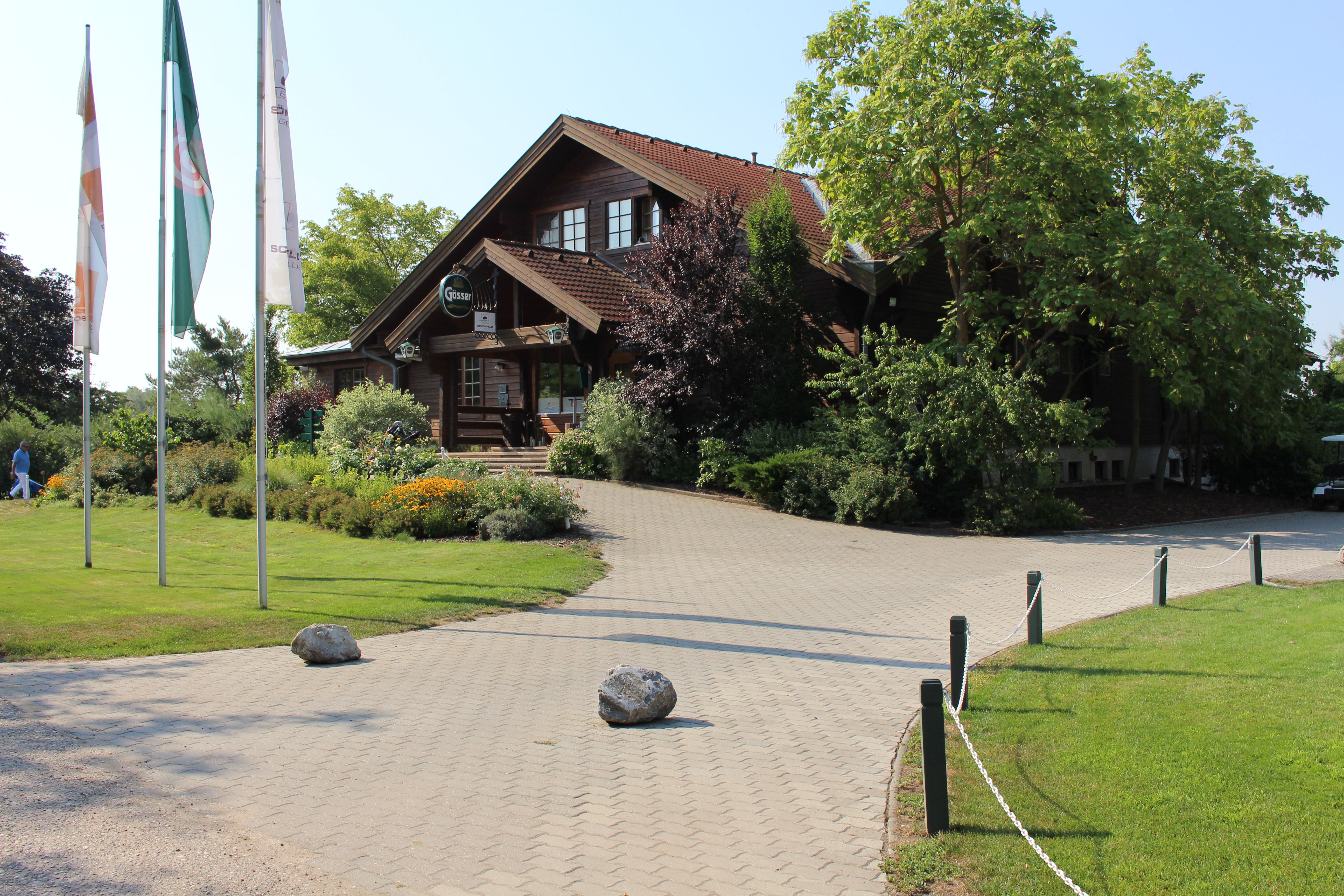 Vchod do golfového klubu s dřevěnou budovou a vlajkami.