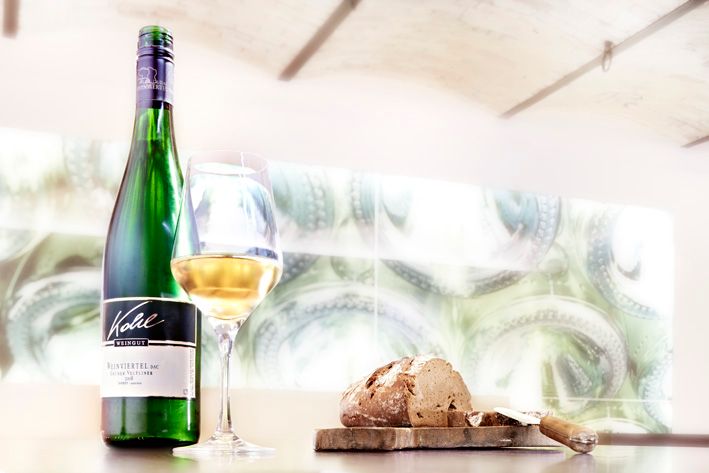 Láhev bílého vína, naplněná sklenička a bochník chleba na stole.