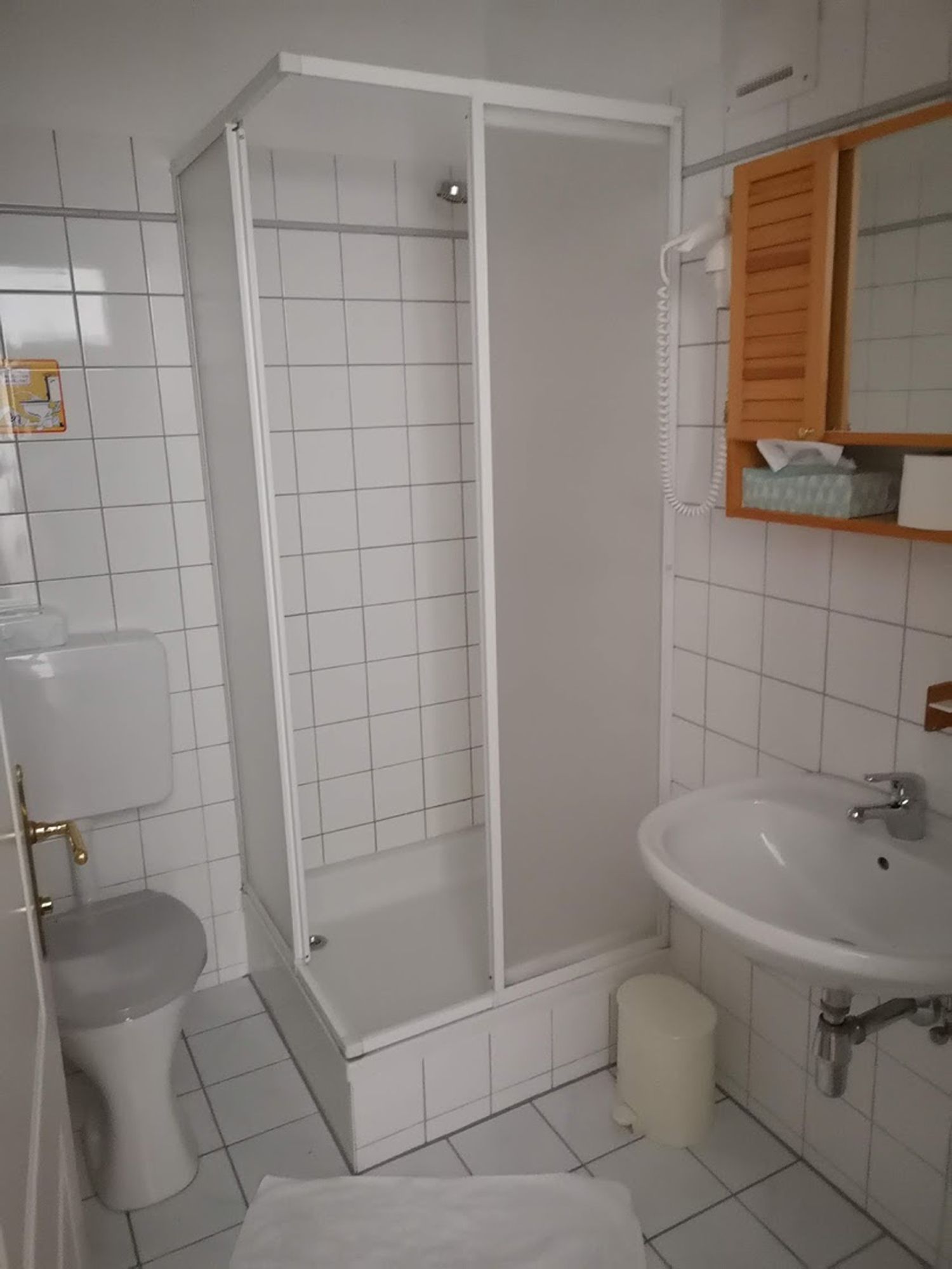 Malá bíle obložená koupelna se sprchovým koutem, toaletou a umyvadlem.