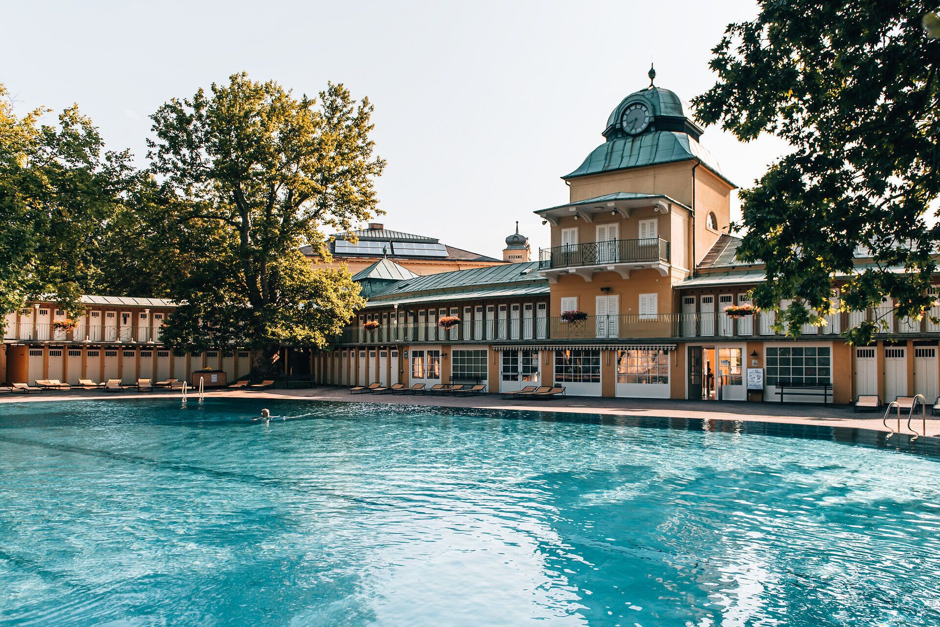 Umgeben von üppigem Grün und majestätischen Bäumen lädt das Thermalbad Vöslau zu erholsamen Stunden ein. Das glitzernde Wasser des Pools reflektiert die warmen Sonnenstrahlen und schafft eine einladende Atmosphäre für Entspannung und Genuss.