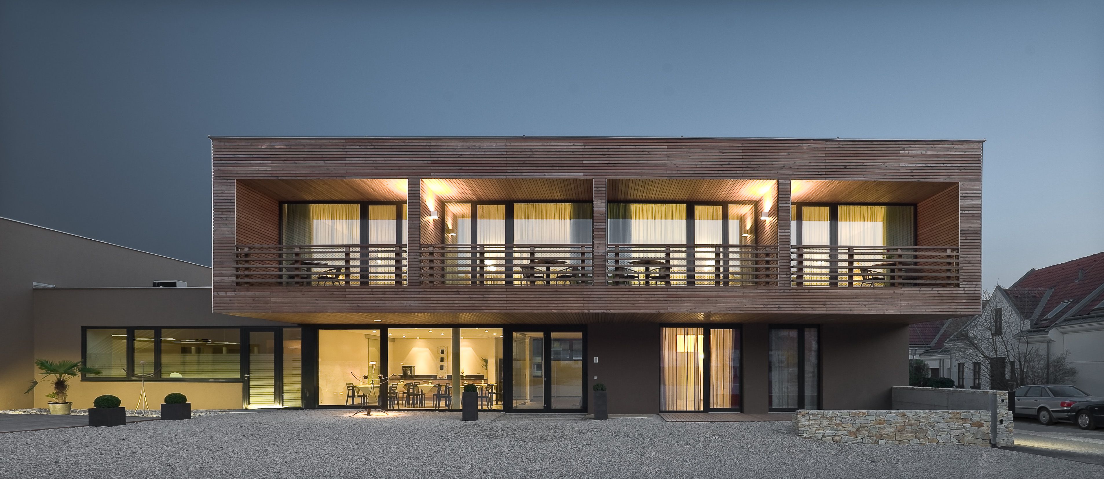 Moderní dvoupodlažní dům s dřevěným obložením a velkými okny s nočním osvětlením.