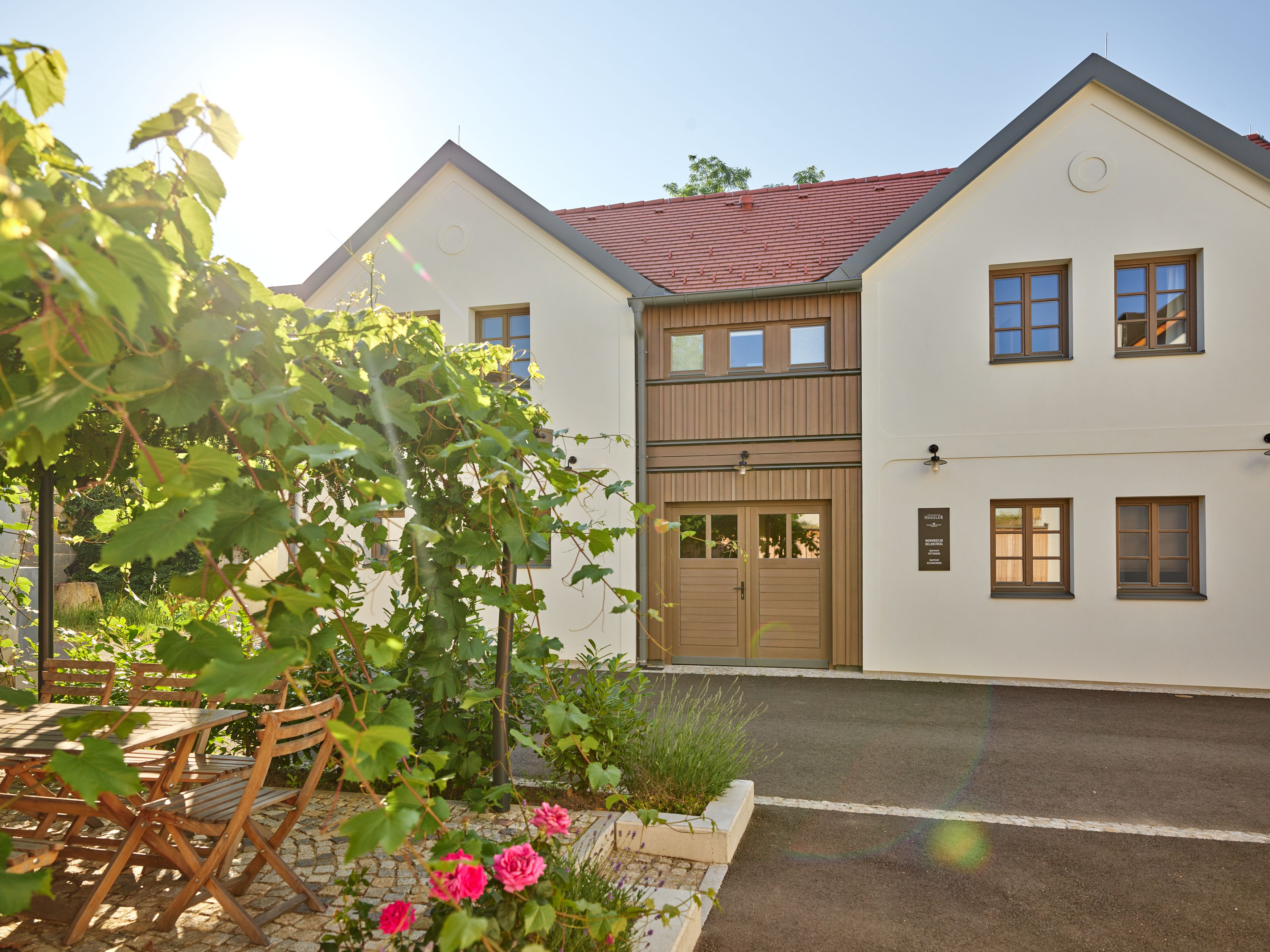 Moderní dvoupodlažní budova s bílými zdmi a červenými střešními taškami. V popředí zahrada s dřevěným stolem, židlemi a kvetoucími rostlinami.