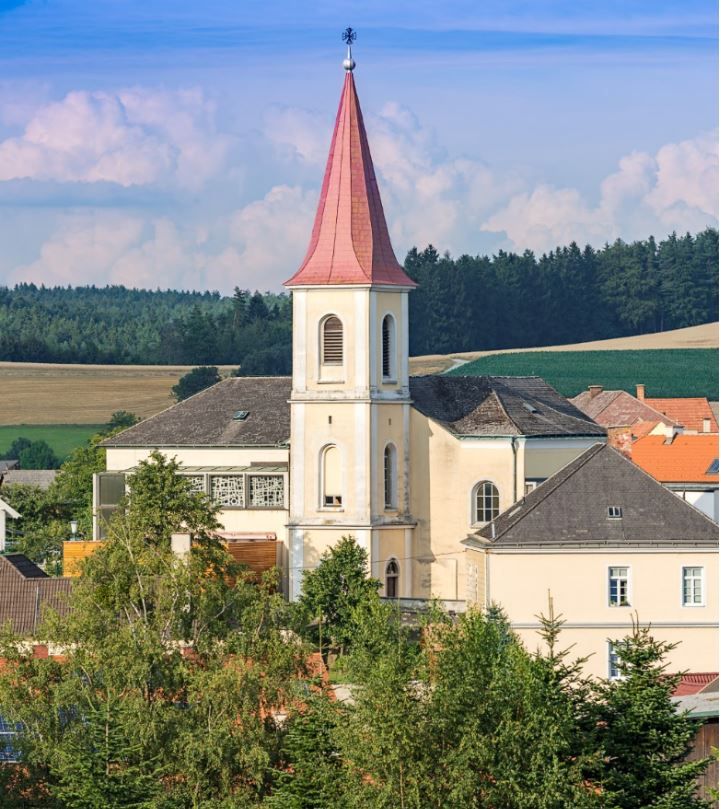 Farní kostel v Hochwolkersdorfu s červenou špičatou střechou věže a okolní krajinou.