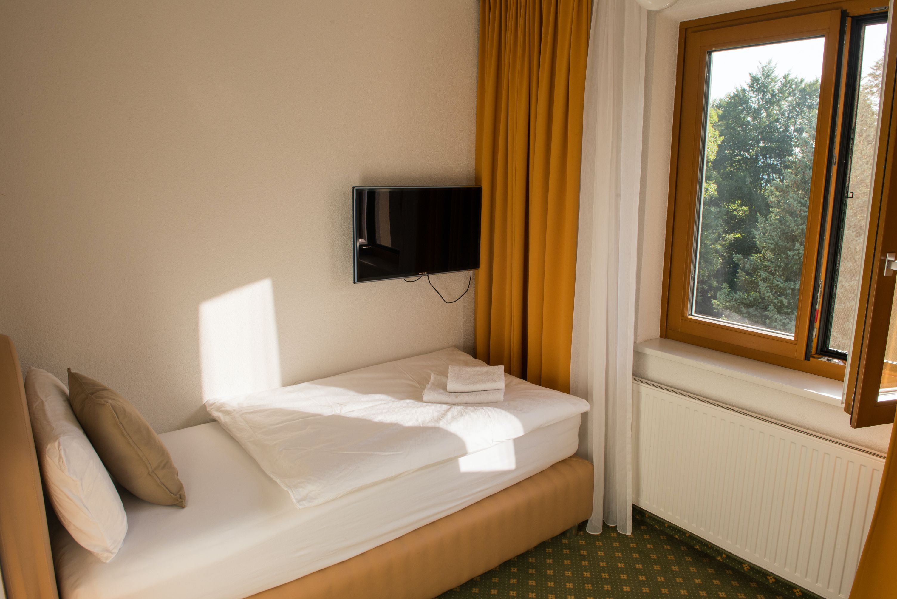 Jednolůžkový pokoj s jednou postelí, žlutými závěsy, oknem s výhledem do zeleně a nástěnnou TV.