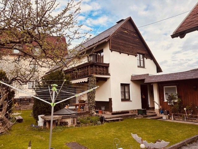 Dvoupodlažní dům s balkonem a zahradou, obklopený stromy a sušárnou v popředí.