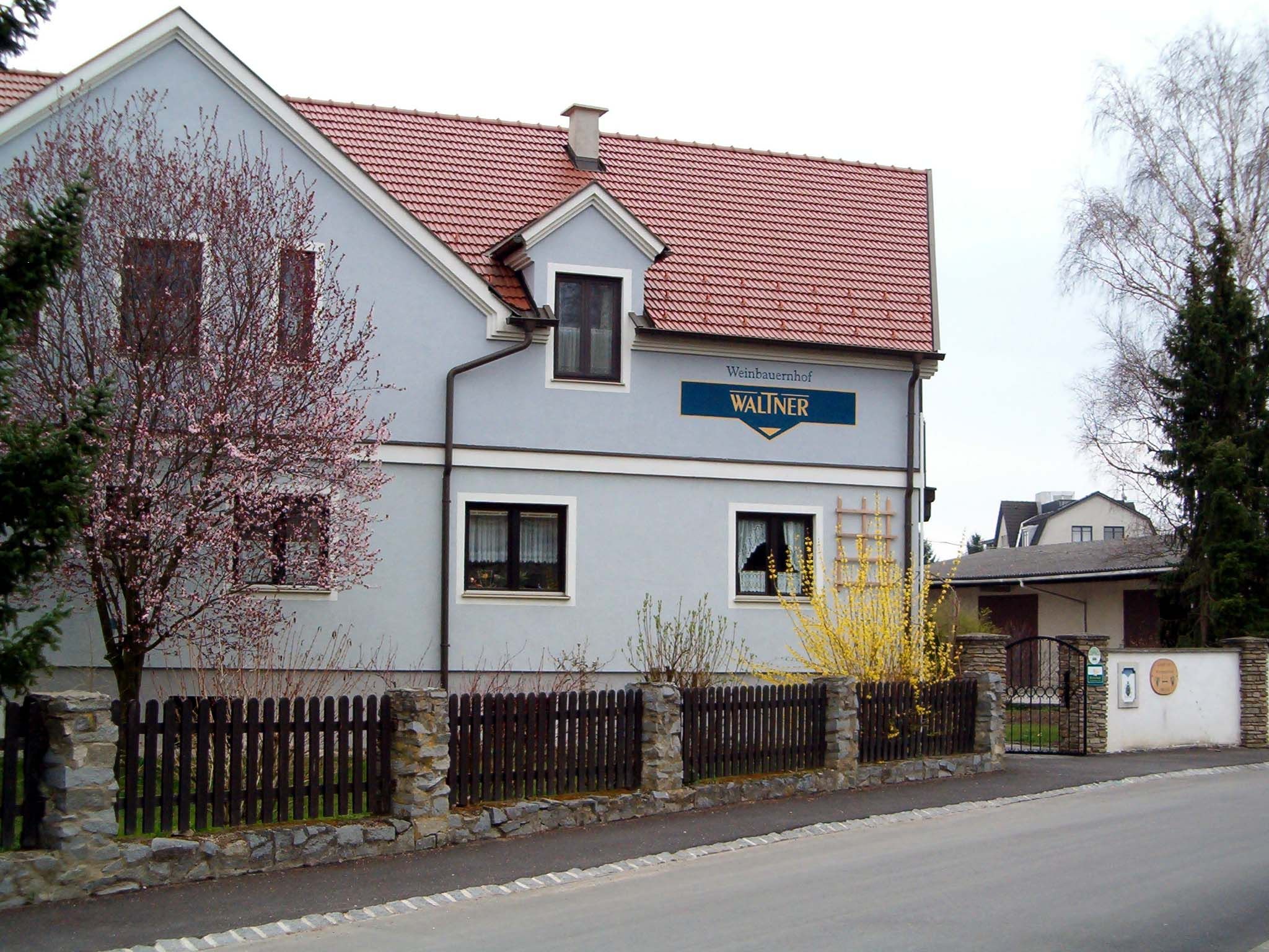 Dvoupatrový dům s červenou střechou a modrou fasádou, obklopený kvetoucími stromy a dřevěným plotem.