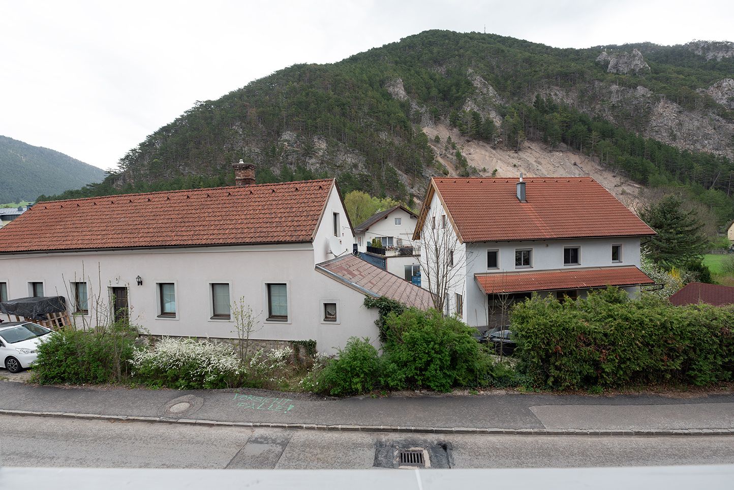 Dva domy s červenými střechami před zalesněným kopcem.