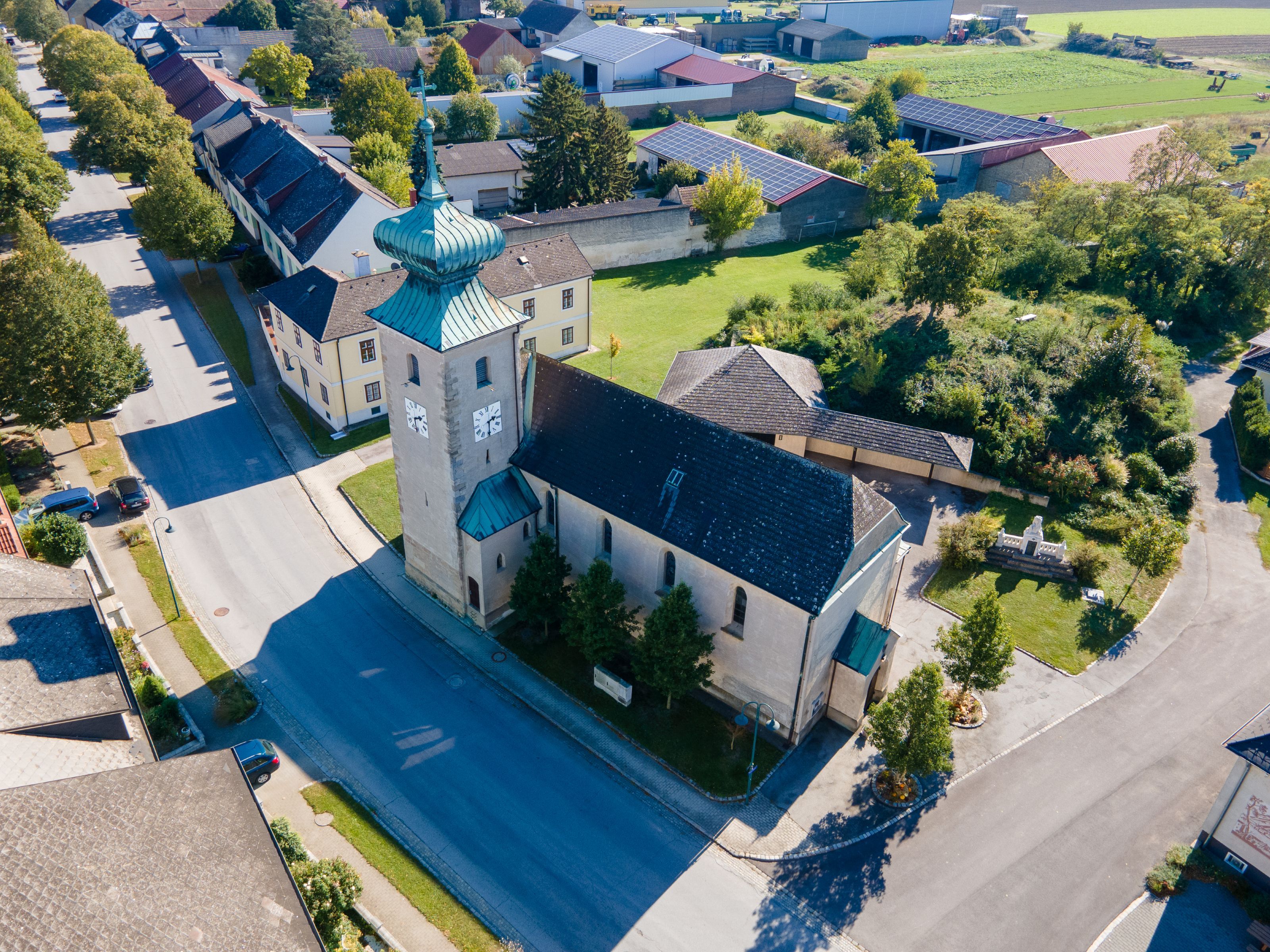 Letecký snímek kostela v obci Weiden an der March s věží, měděnou střechou a okolními domy a zahradami.