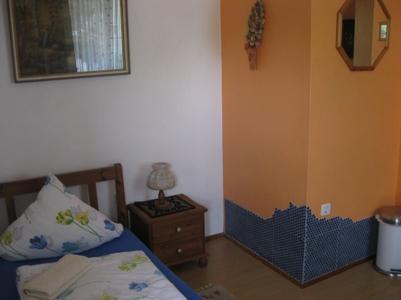 Malý pokoj s jednou postelí, nočním stolkem, lampičkou a oranžovou stěnou s mozaikovými dlaždicemi.