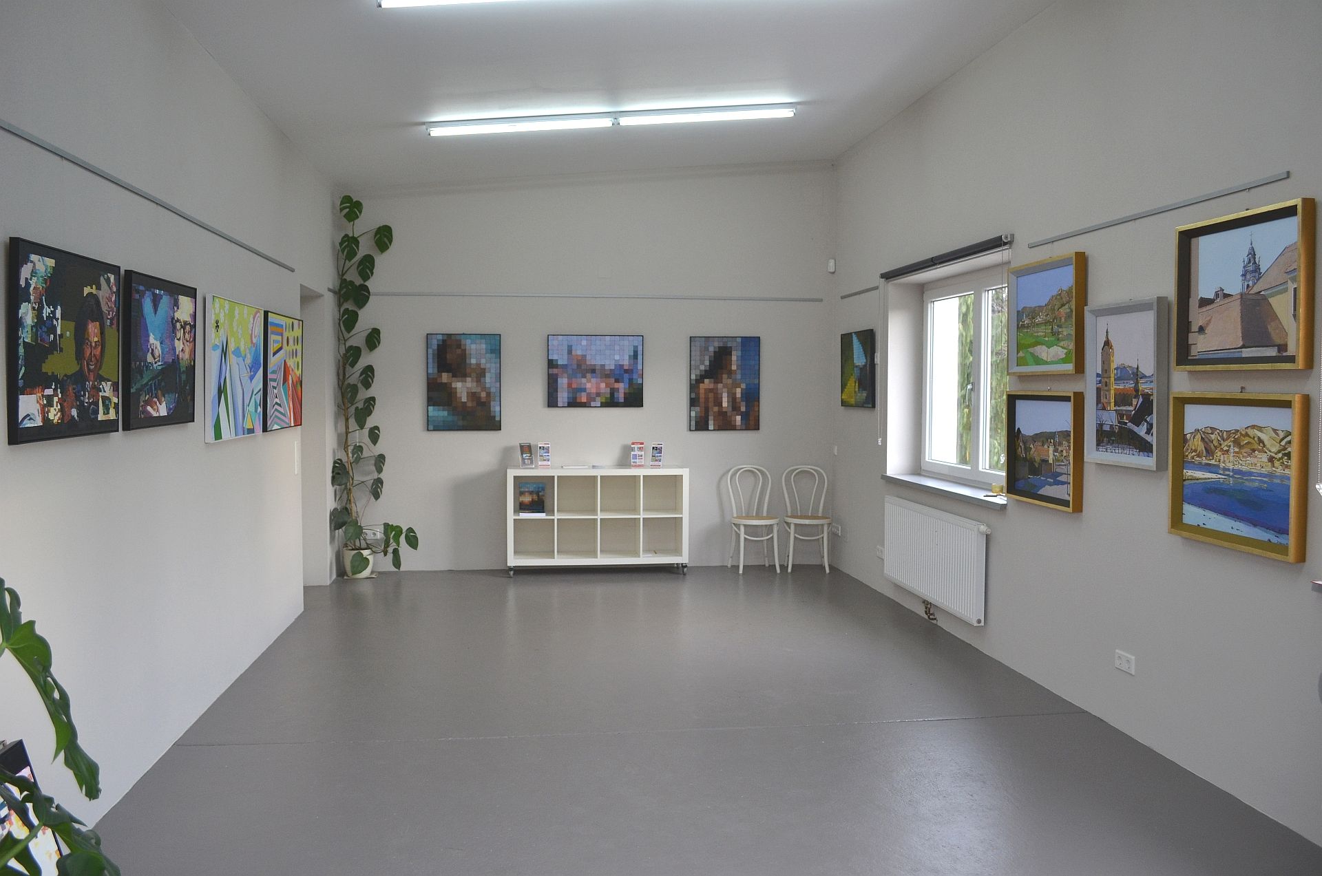 Pohled do interiéru galerie s obrazy na stěnách a policí uprostřed.