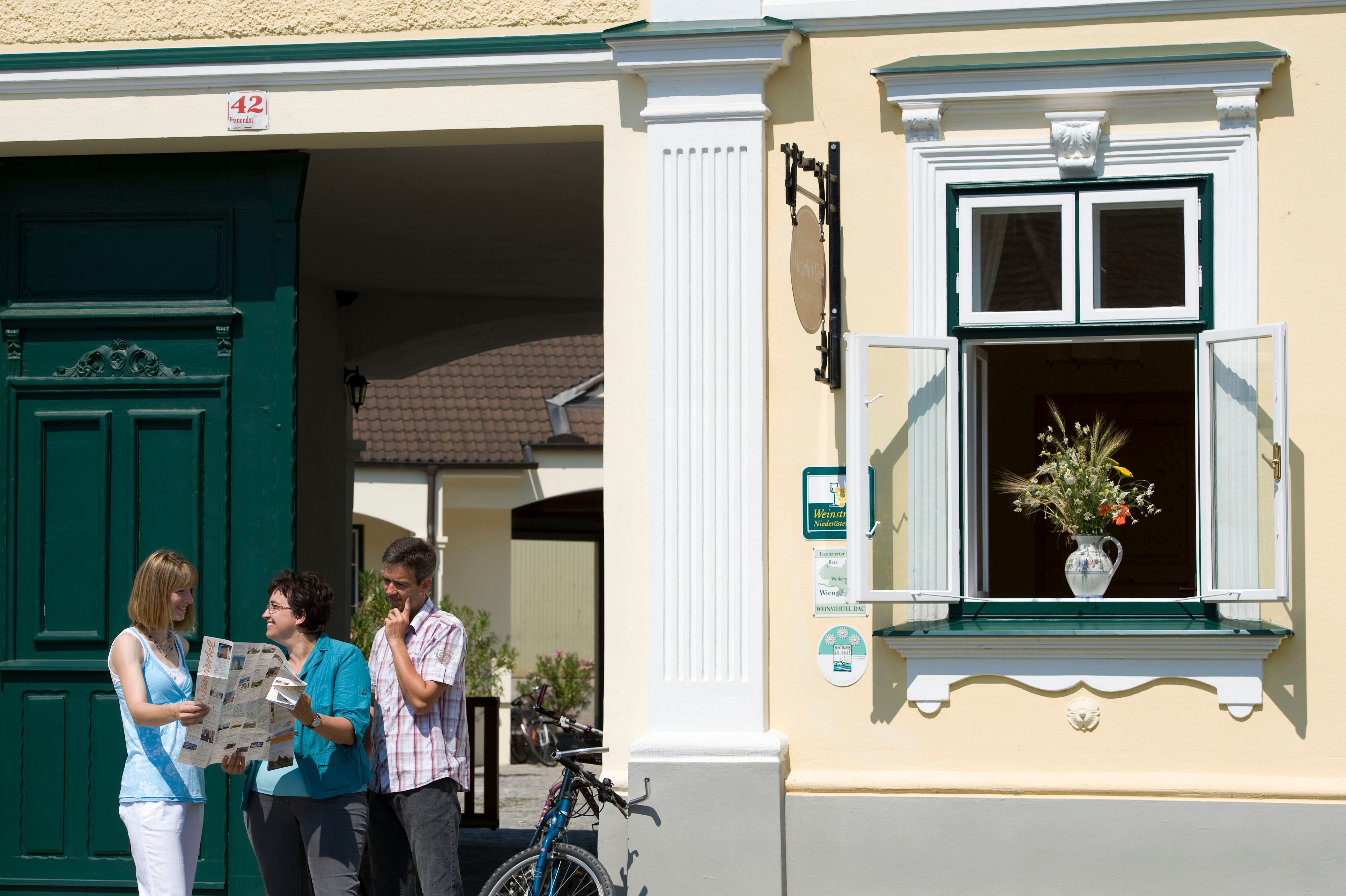 Před žlutou budovou se zelenými dveřmi stojí tři lidé a čtou si mapu. Okno s kyticí květin je otevřené.