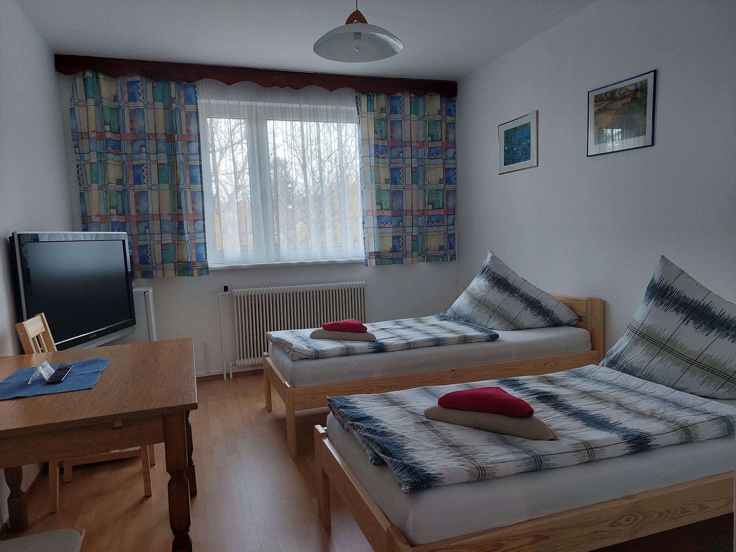 Jednoduše zařízený pokoj se dvěma samostatnými lůžky, stolem, TV a oknem s barevnými závěsy.