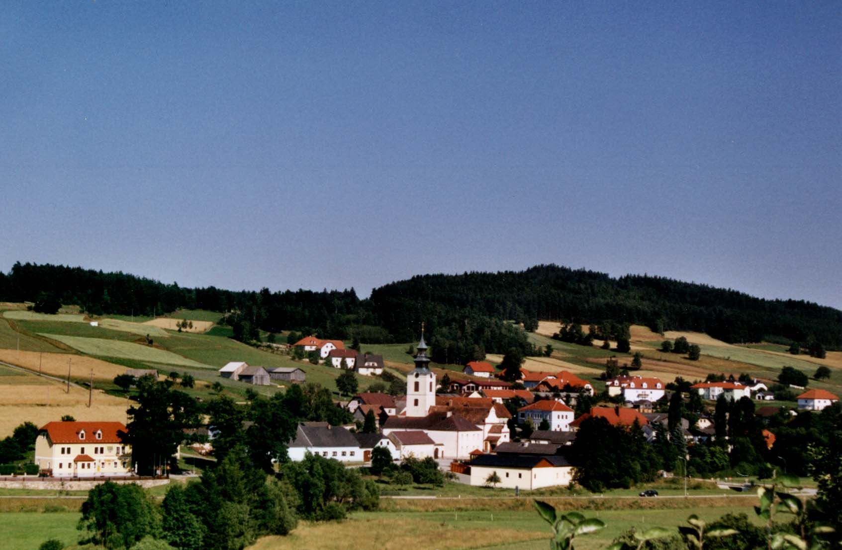 Panorama vesnice s kostelem a červenými střechami, obklopené zelenými poli a lesy.