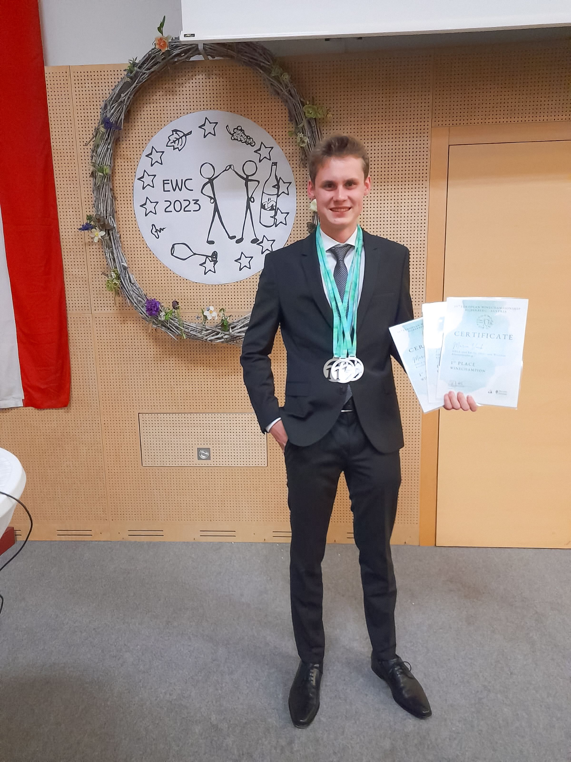 Mladý muž v obleku stojí před ozdobným věncem s certifikátem a medailemi v ruce.