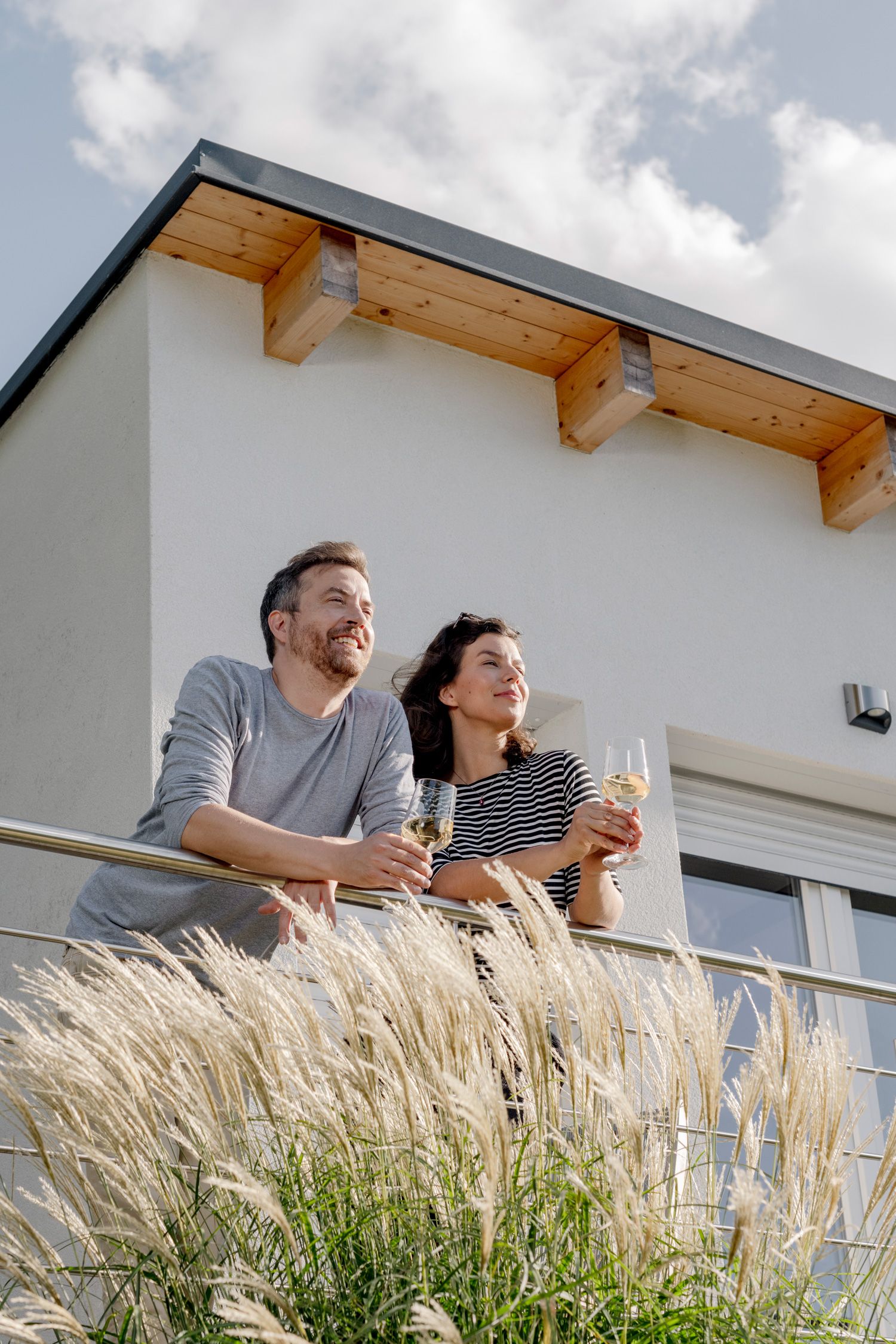 Pár stojí na balkoně se sklenicemi vína a v pozadí je moderní budova.