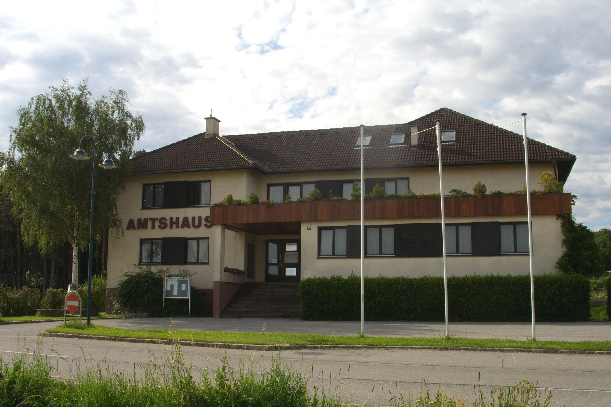 Kancelářská budova Natschbach-Loipersbach s hnědou střechou a balkonem, obklopená stromy a stožáry.