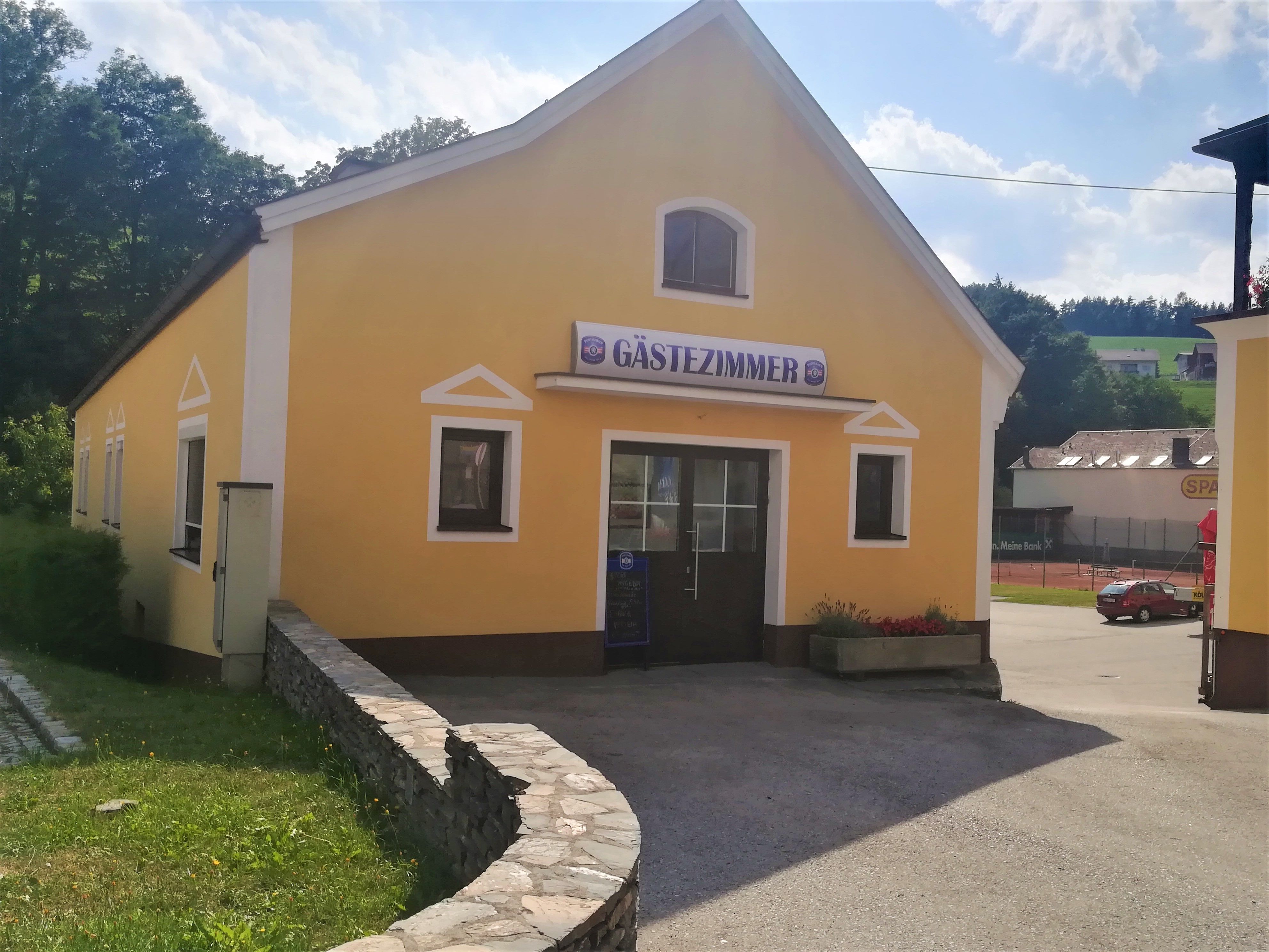 Žlutá budova s nápisem "guest room", obklopená stromy a parkovištěm.