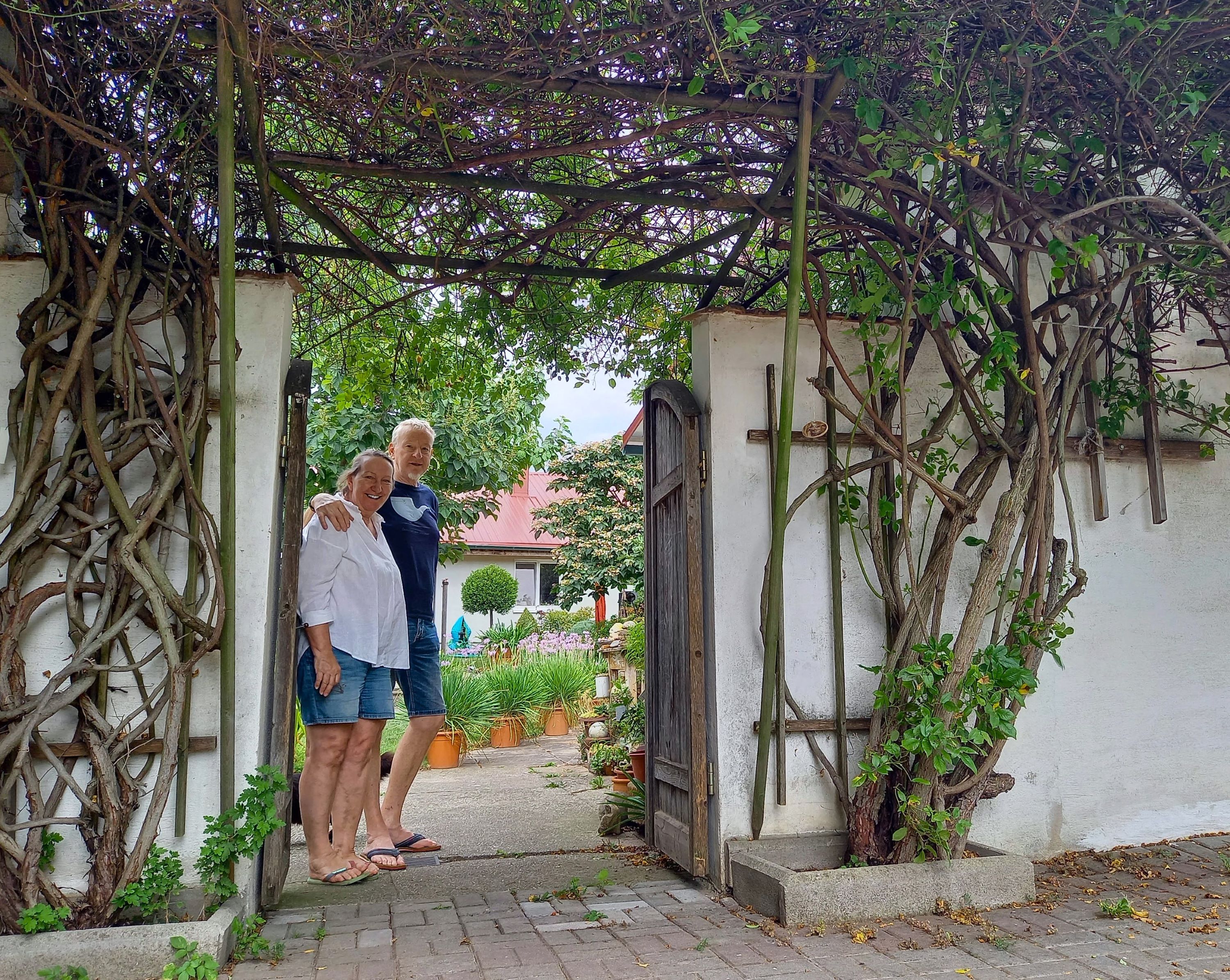 Dva lidé stojí s úsměvem pod obloukem zarostlým rostlinami v zahradě.